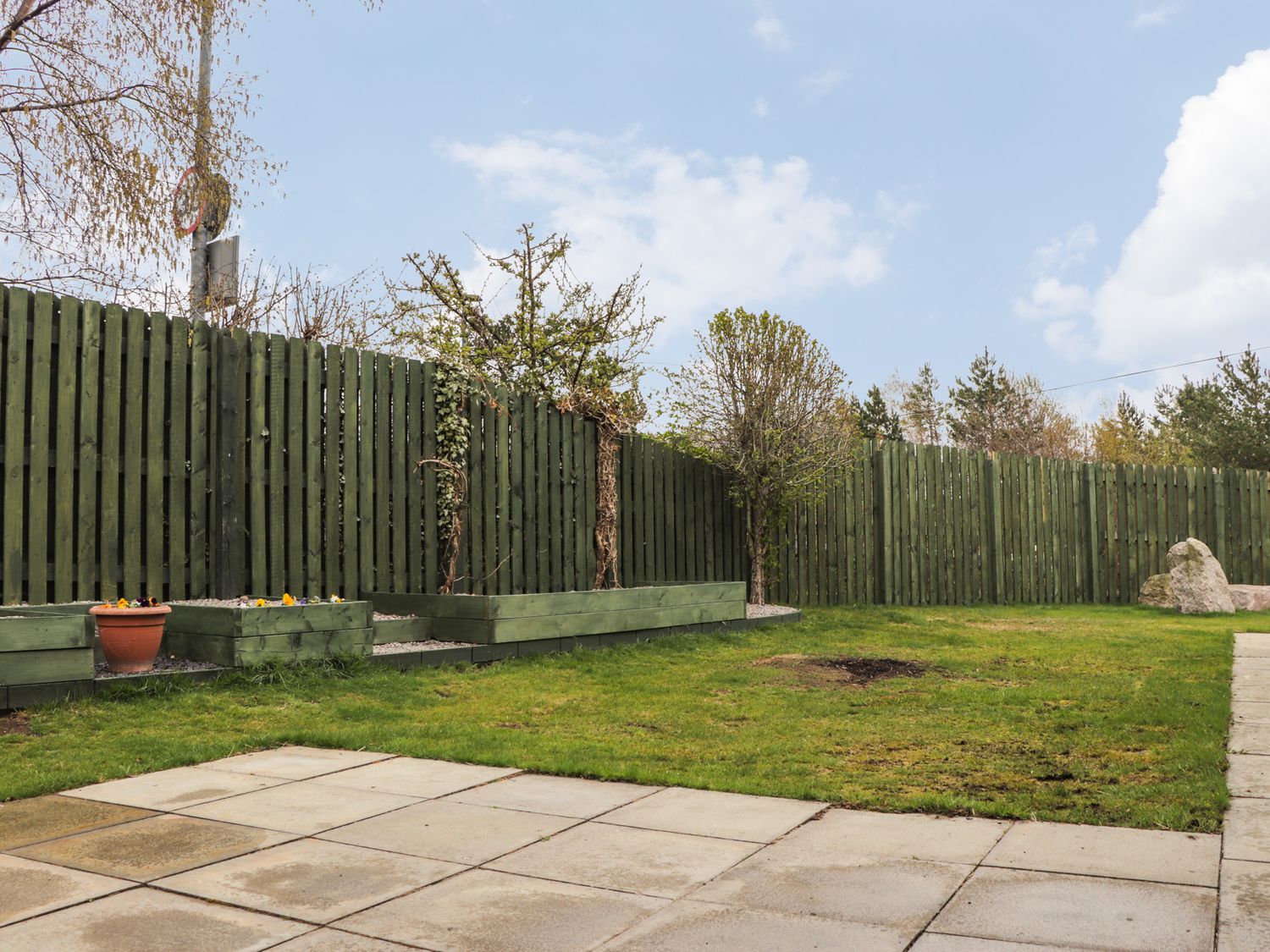 A fenced backyard with green grass, paved patio area, small trees, flower planters and rocks at 3 Croftside in Aviemore