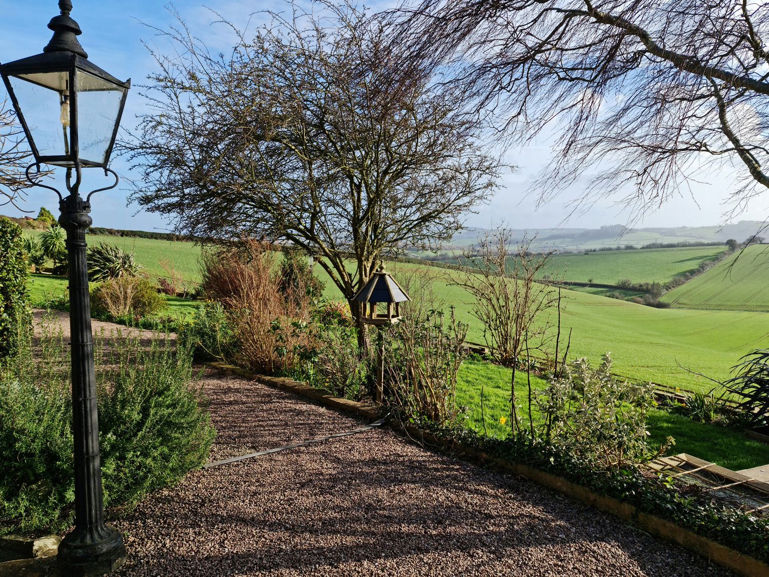 A garden with a gravel path lamp post bird feeder trees and bushes overlooking green fields at The Thatched Cottage in Crediton