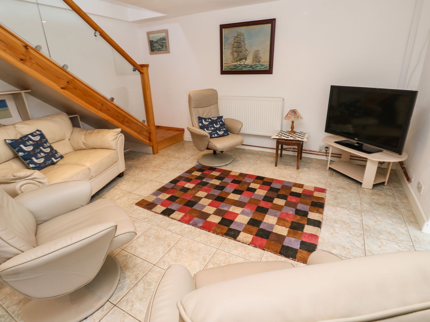 A sitting room with beige armchairs a sofa a multicolored checkered rug a television on a stand and a wooden staircase at Alice Cottage in Wheal Kitty near St Agnes