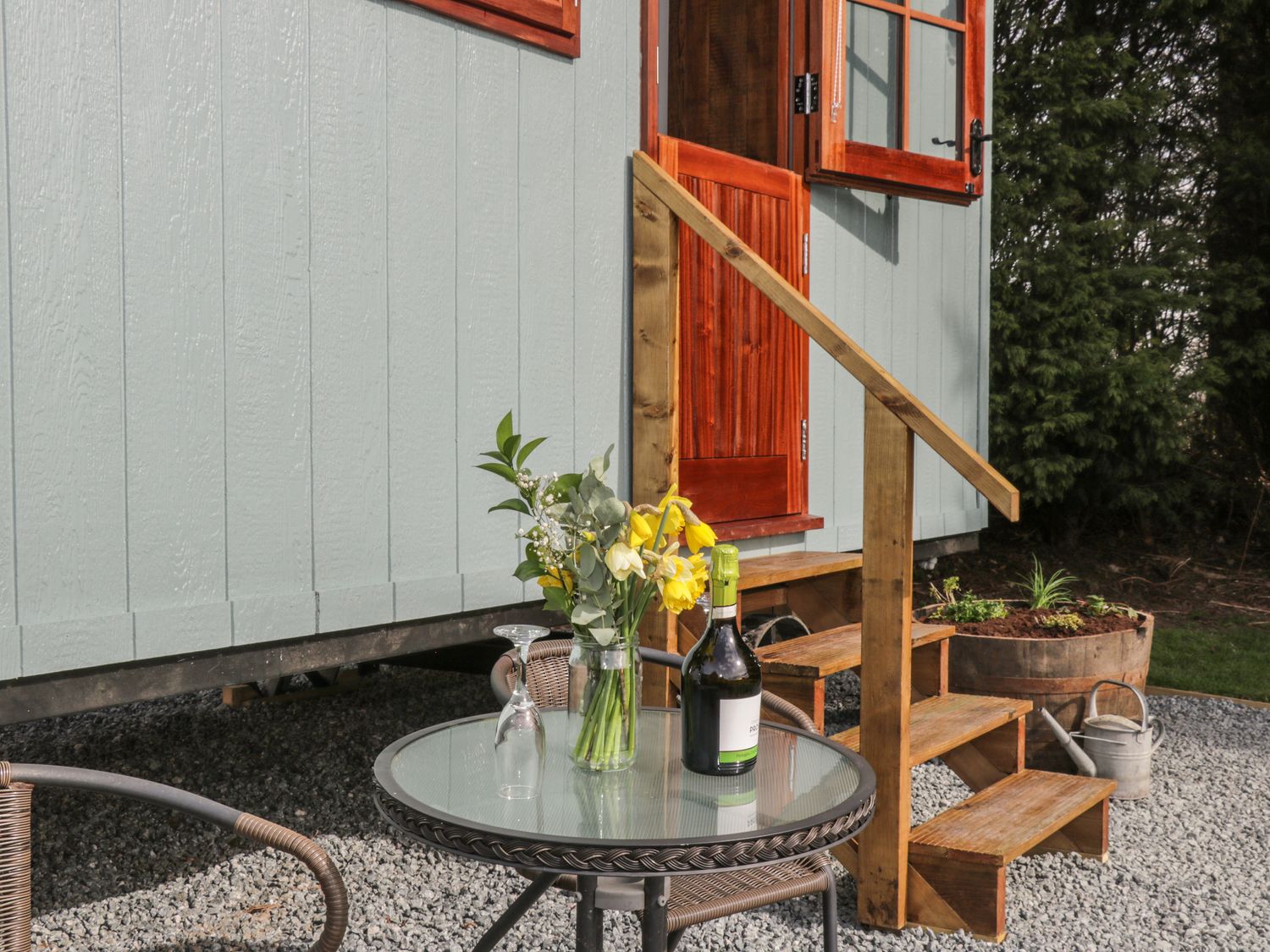 An outdoor seating area with a glass top table holding flowers a wine bottle and an upside down glass near wooden steps and a door at Ewes Welcome in Cottingham