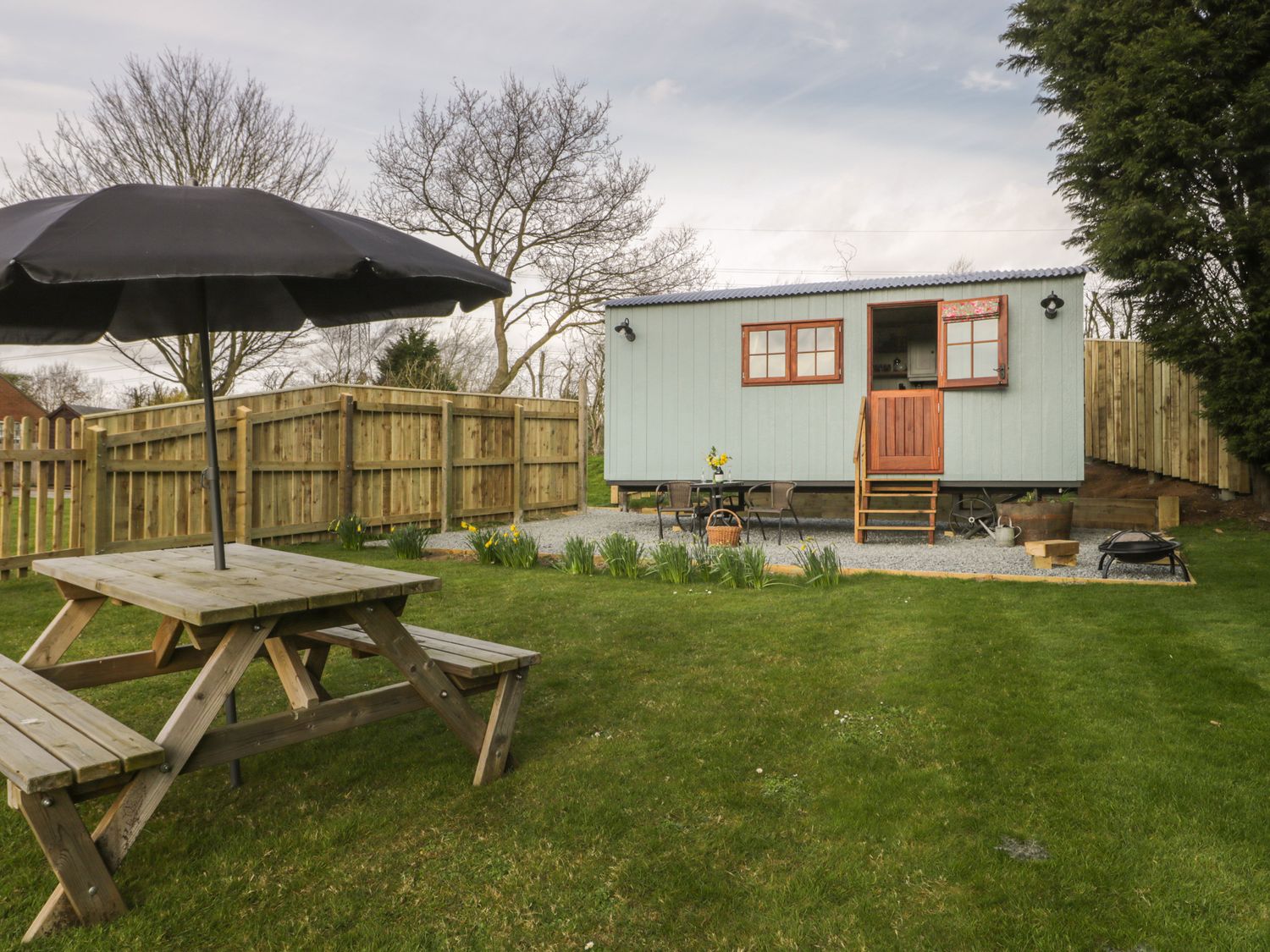 A fenced garden with a wooden picnic table and umbrella and a small light blue cabin with stairs at Ewes Welcome in Cottingham