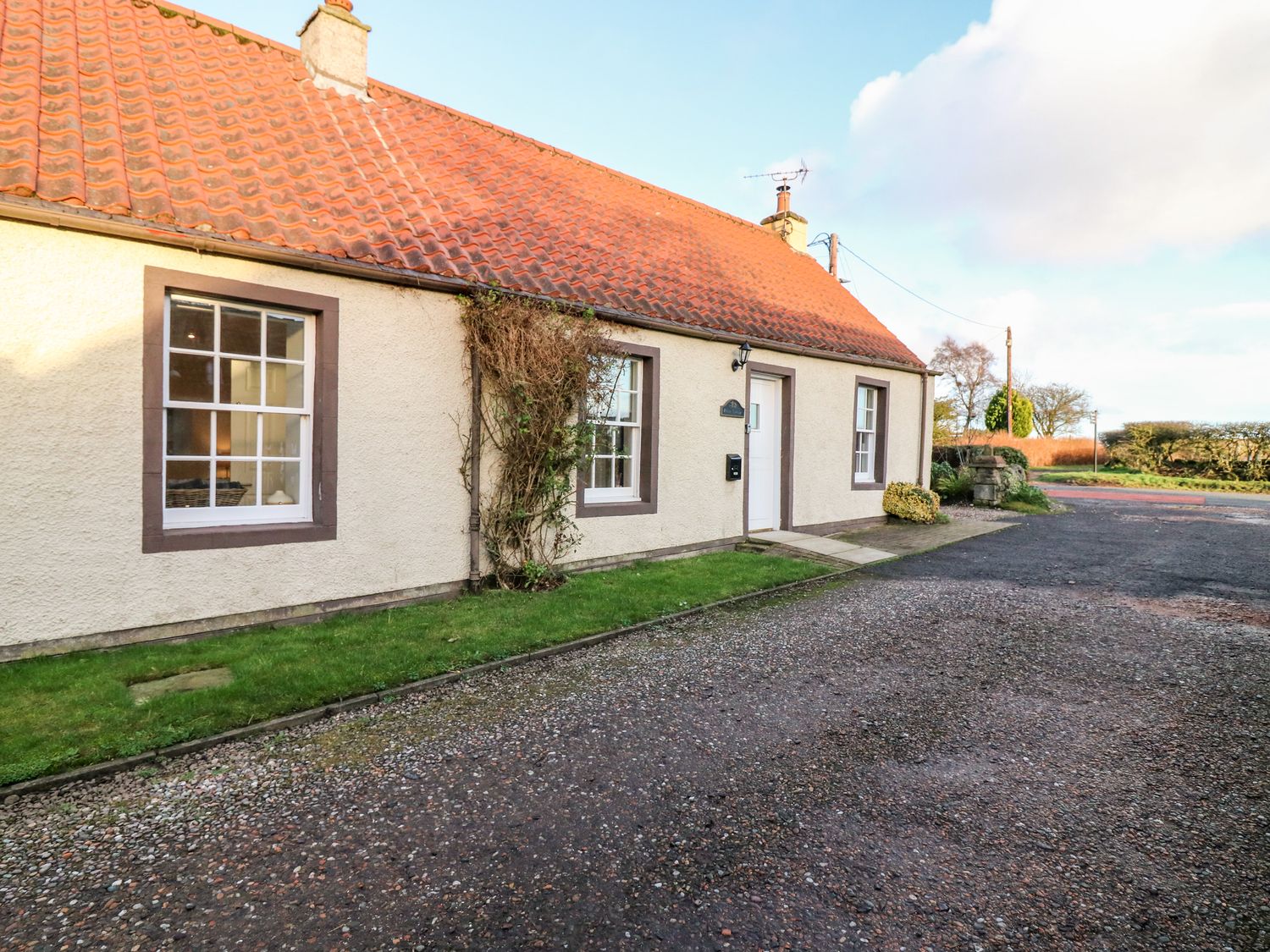 The exterior of a single-story house with a tiled roof grass along the front and a gravel driveway at Rhum Cottage Peat Inn near St Andrews