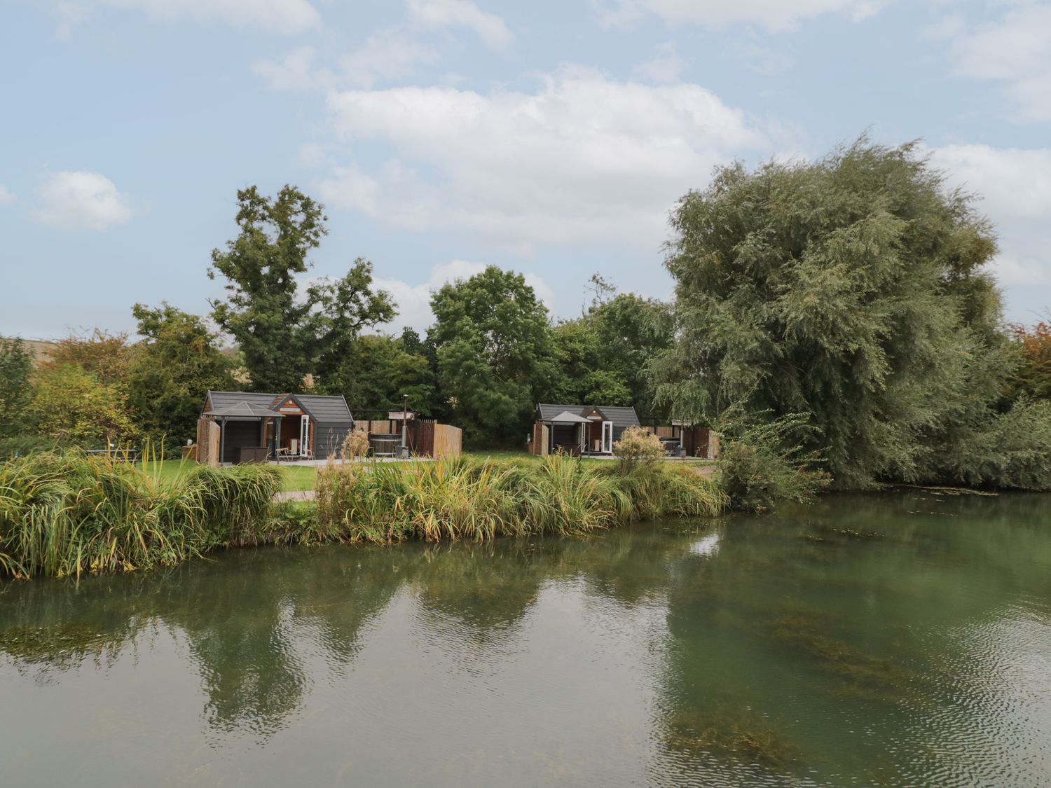 Two small wooden cabins near a pond with grass and trees at Willow Lodge At Bridge Lake Farm & Fishery in Chacombe