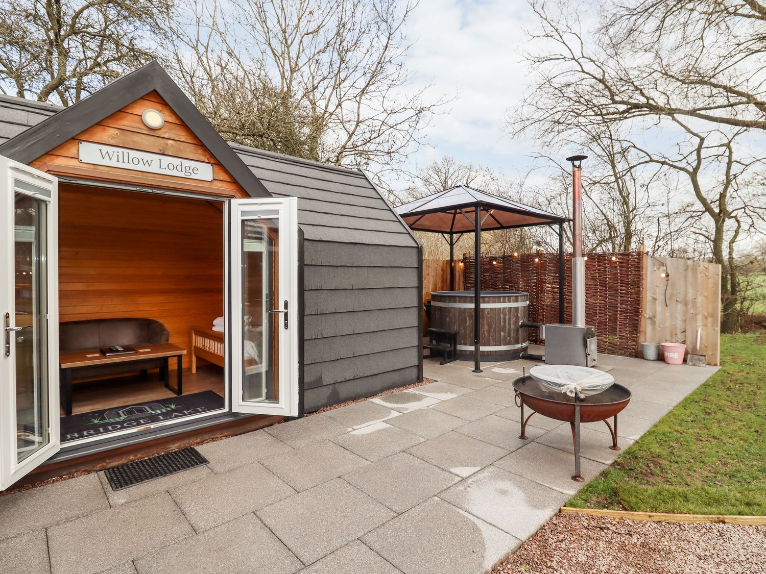 An outdoor patio with a wooden cabin labeled Willow Lodge a hot tub under a gazebo and a fire pit at Willow Lodge At Bridge Lake Farm & Fishery in Chacombe
