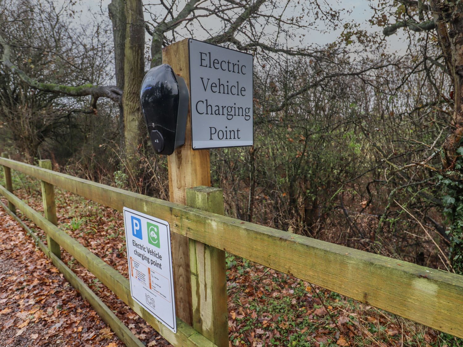 An electric vehicle charging point mounted on a wooden post next to a wooden fence with trees in the background at Willow Lodge At Bridge Lake Farm & Fishery in Chacombe