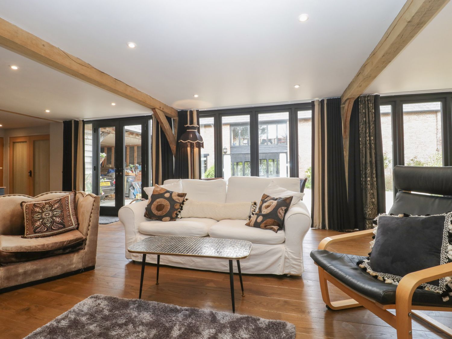 A living room with a white sofa, armchairs, a coffee table, wooden beams, and large windows at The Courtyard - Hilltop Barn in Winterborne Zelston near Wareham