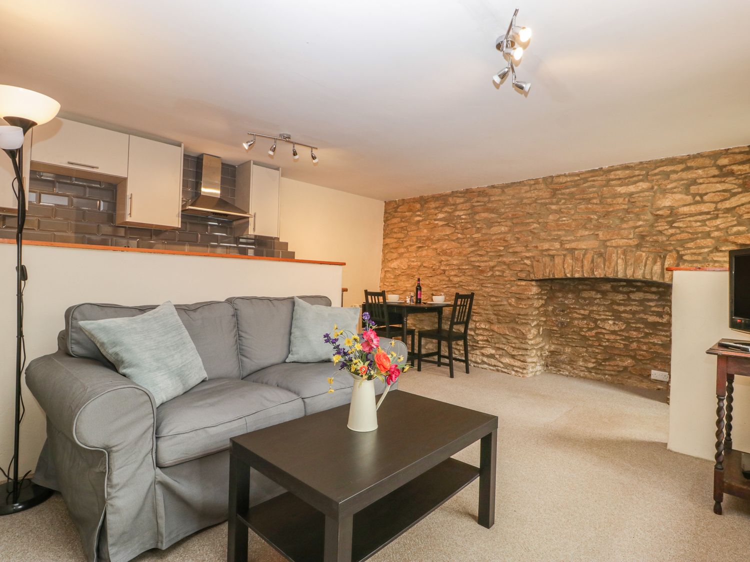 A living room with a gray sofa and cushions a black coffee table with a flower vase a dining table with chairs against a stone wall and a kitchen area with white cabinets and a stainless steel hood at Jasmine Cottage in South Perrott near Mosterton