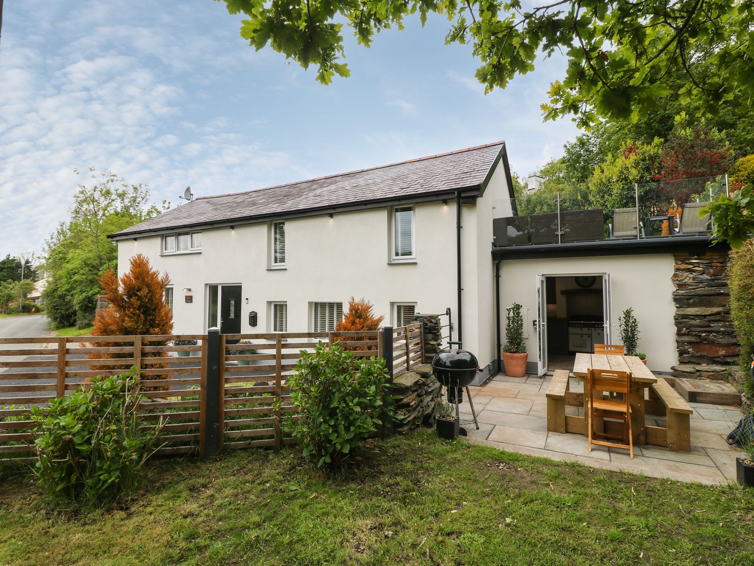 An outdoor patio area with a wooden picnic table and chairs a barbecue grill and open doors leading into a kitchen at Bwthyn Bach Gwyn in Penrhyndeudraeth