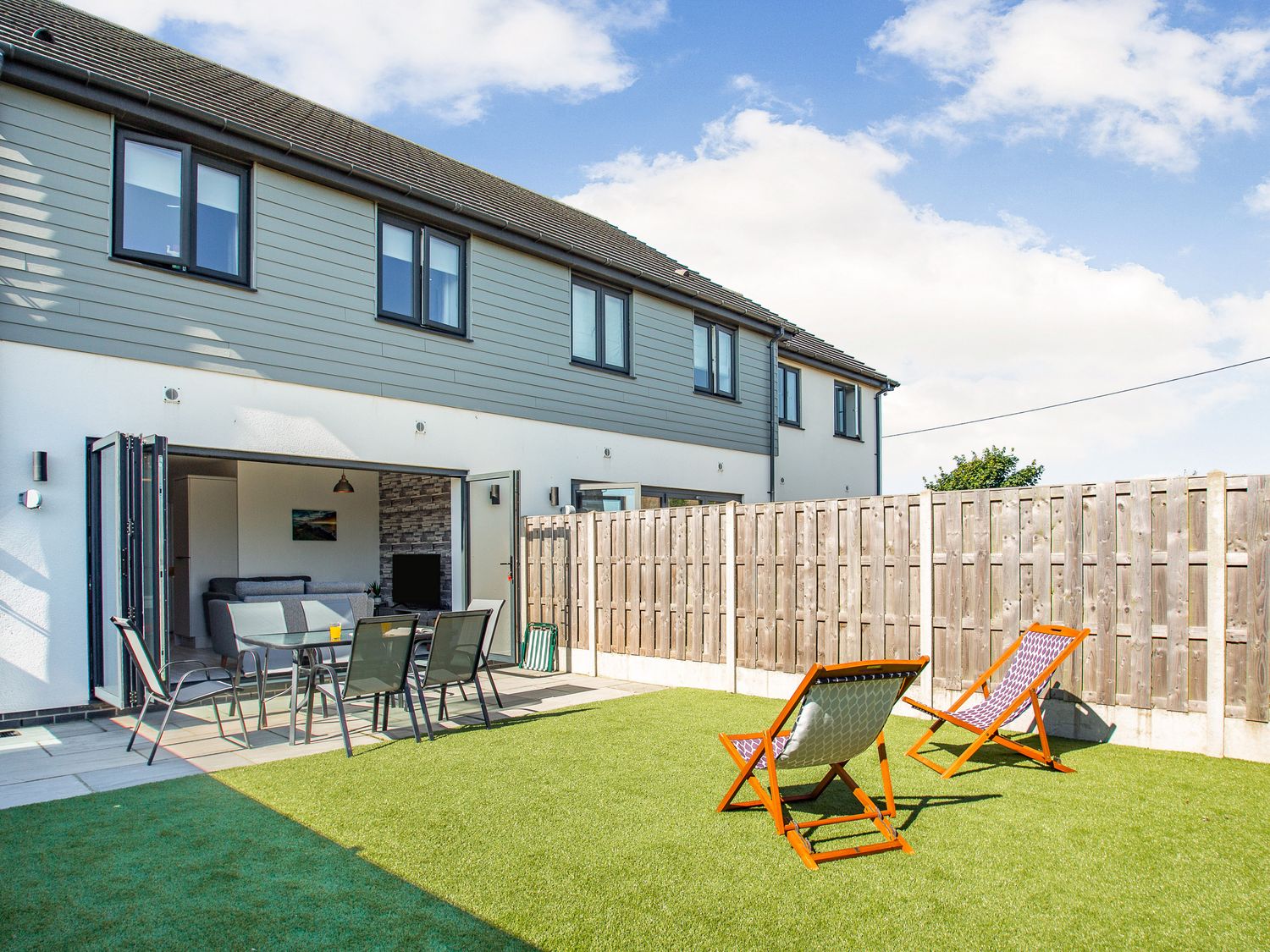A backyard with two deck chairs and a glass table with chairs outside a house with open doors at Sea View in Llanfaelog