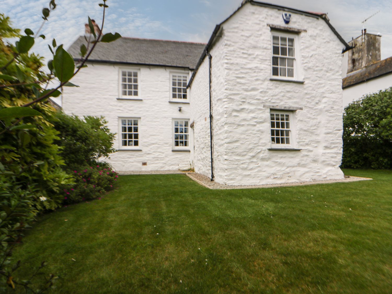 A white stone house with multiple windows and a green lawn surrounded by bushes at Great Western Farmhouse in Crantock