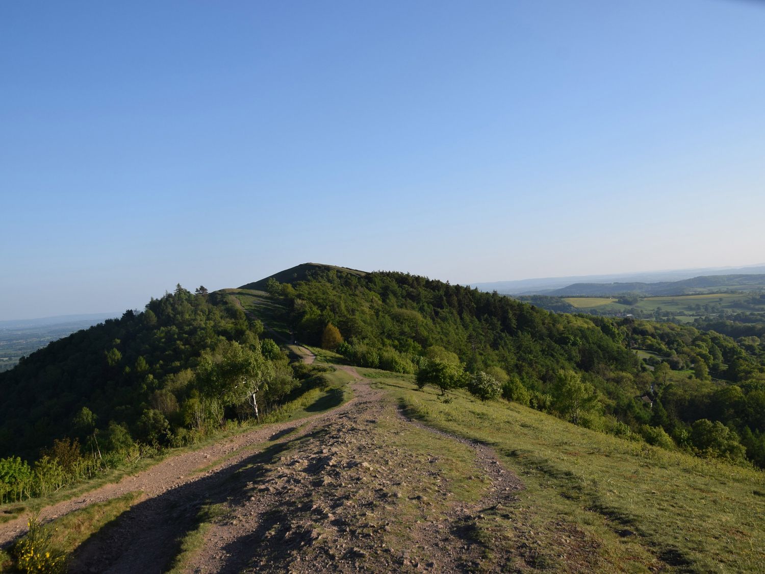 A dirt path running through a grassy hill with trees and a view of distant hills at The Escape in Malvern