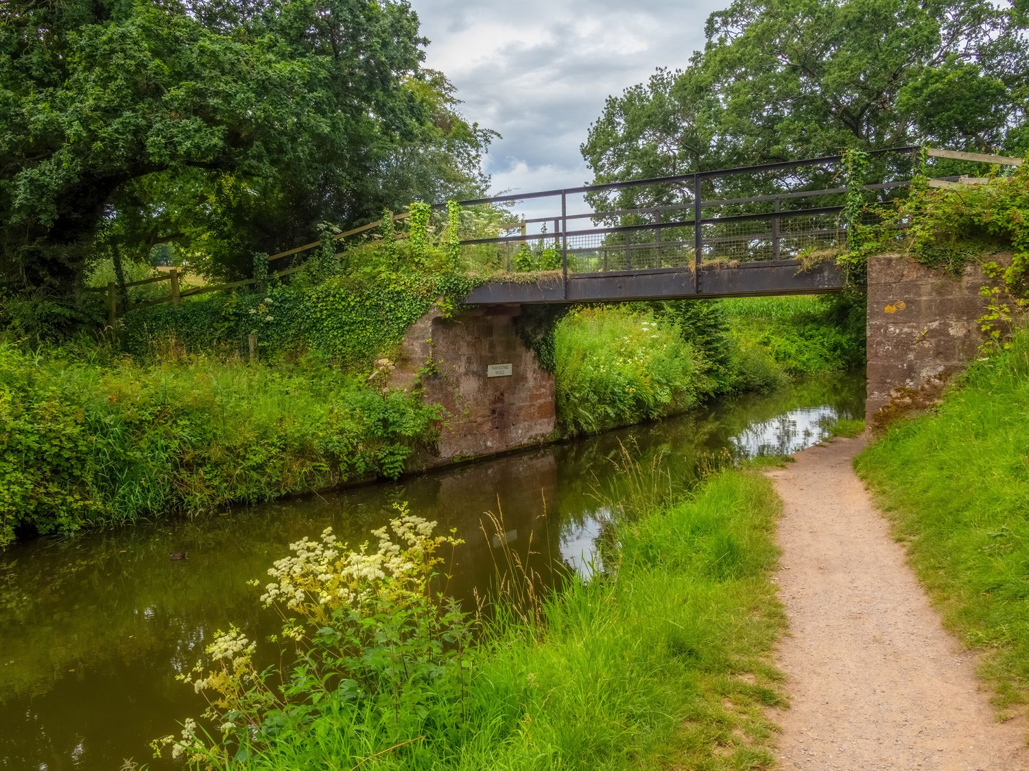 A small canal with a metal bridge over it and a dirt path alongside at The Linhay in Cadeleigh near Tiverton