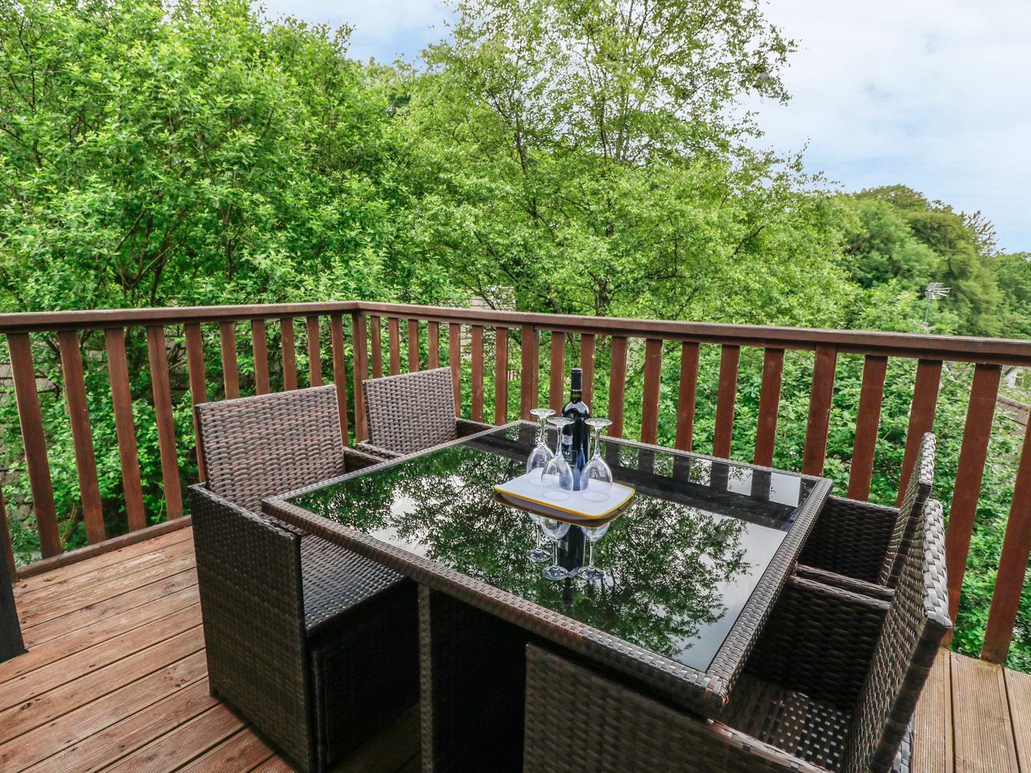 An outdoor wooden deck with a glass top table surrounded by four wicker chairs and a bottle with four wine glasses on the table at 19 Honicombe Park in St Ann's Chapel near Drakewalls