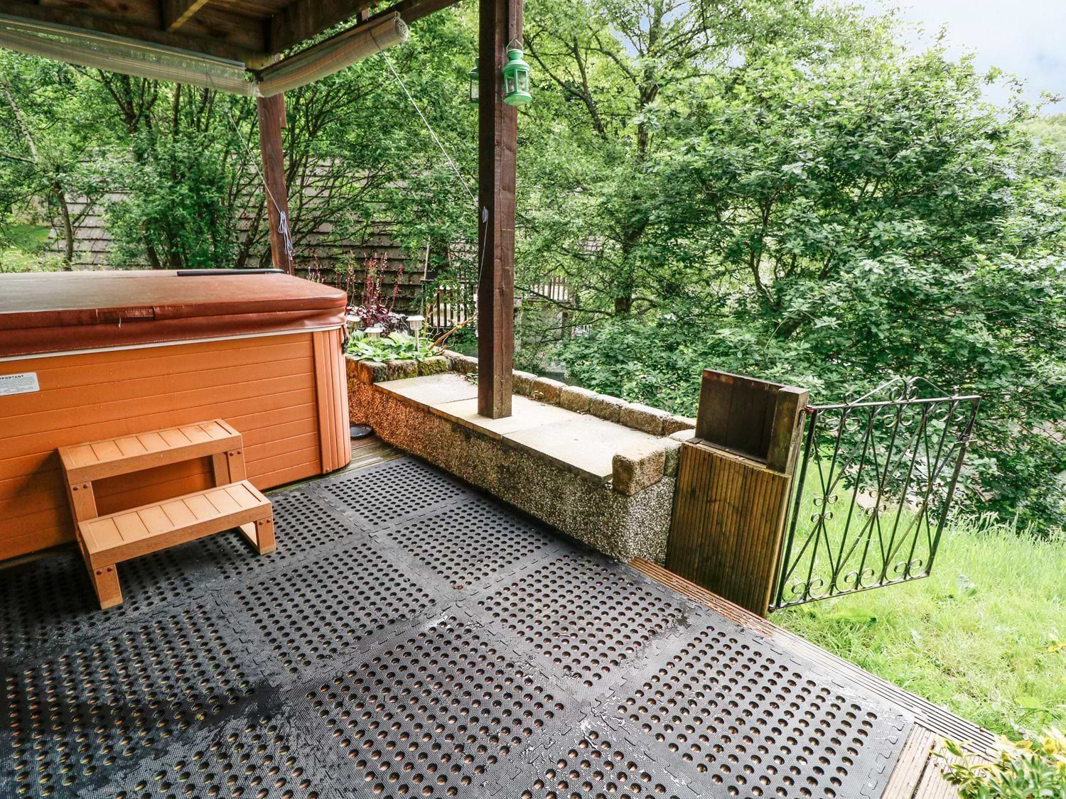 An outdoor covered area with a hot tub wooden steps and a metal gate overlooking grass and trees at 19 Honicombe Park St Ann's Chapel near Drakewalls