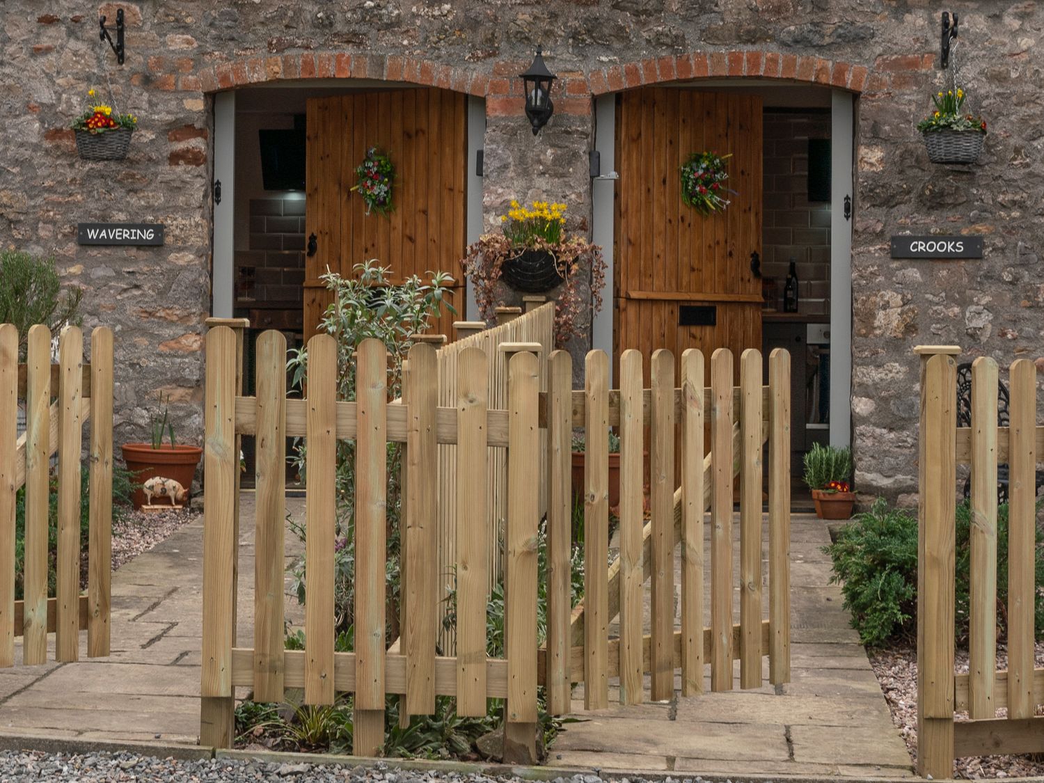 A stone building with two wooden doors labeled wavering and crooks with plants and a wooden fence outside at Crooks in Axbridge