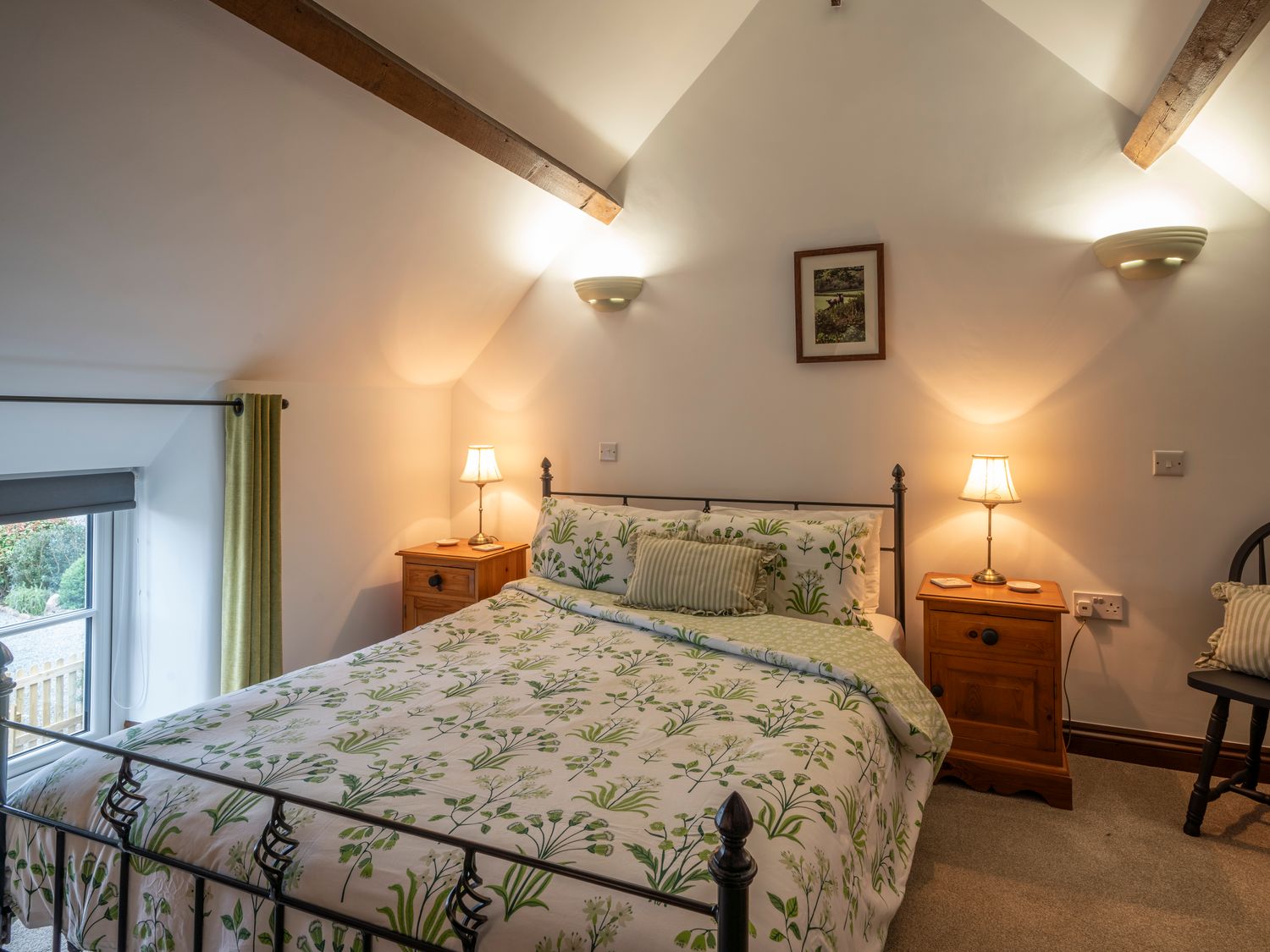 A bedroom with a bed with floral bedding two bedside tables with lamps a window with curtains and wooden beams on the ceiling at Crooks in Axbridge