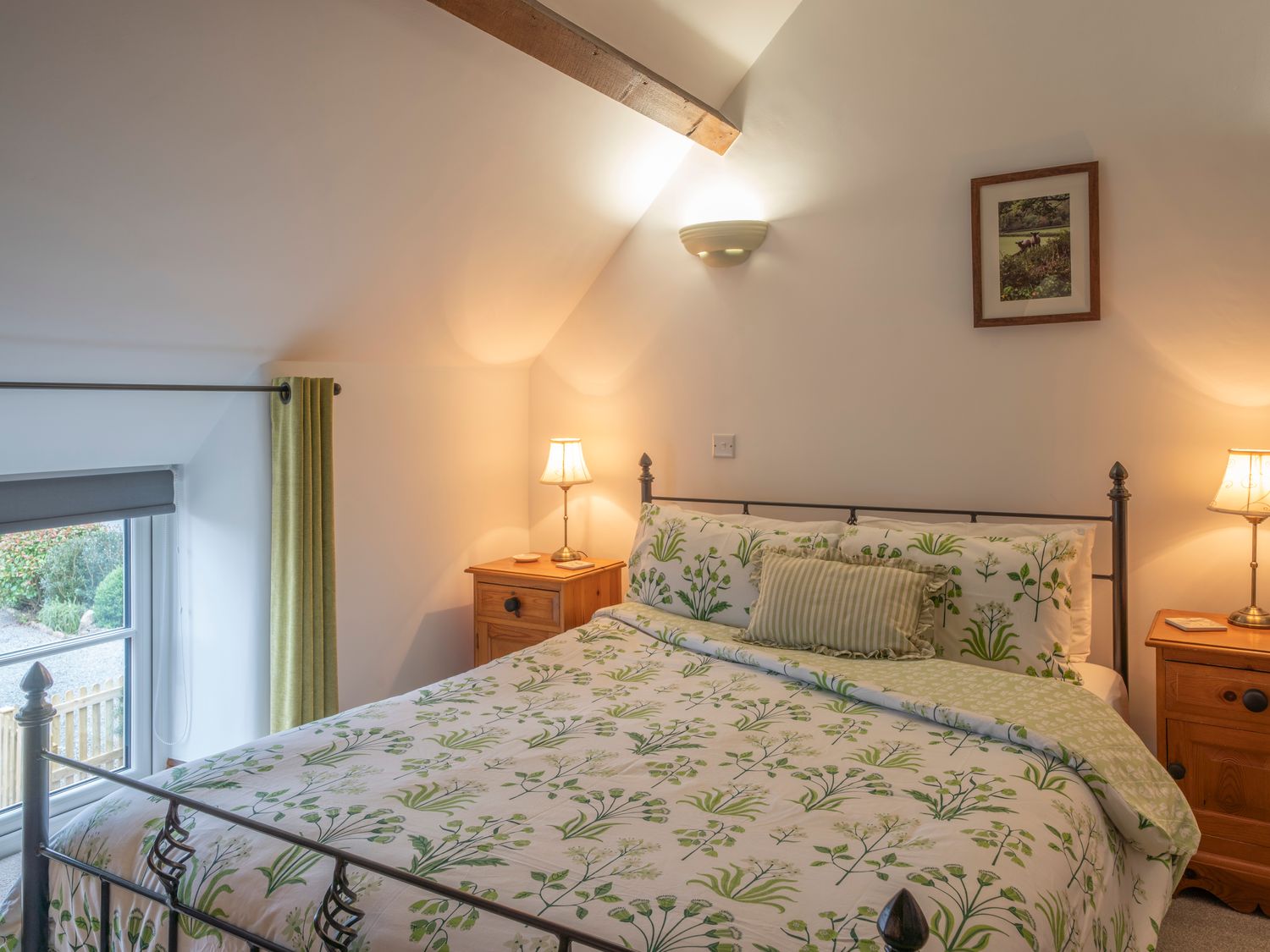 A bedroom with a bed with floral bedding two wooden bedside tables with lamps and a window with green curtains at Crooks in Axbridge