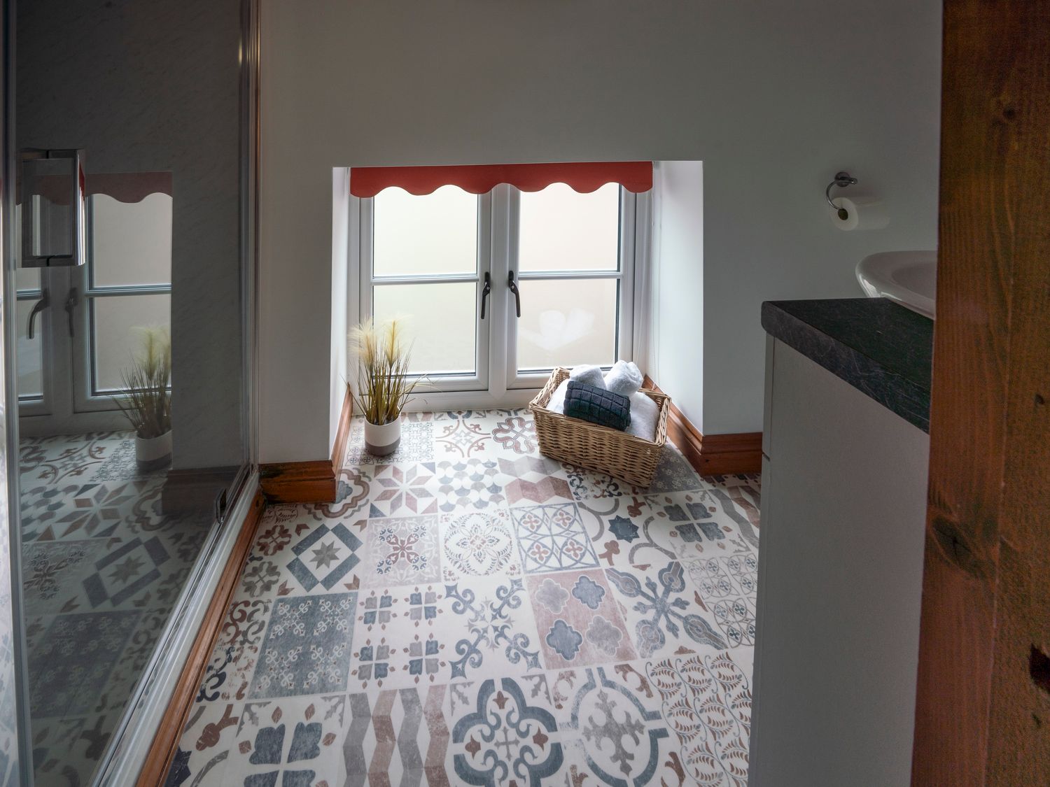 A bathroom with patterned floor tiles a basket with towels a window with a red valance and a potted plant at Crooks in Axbridge