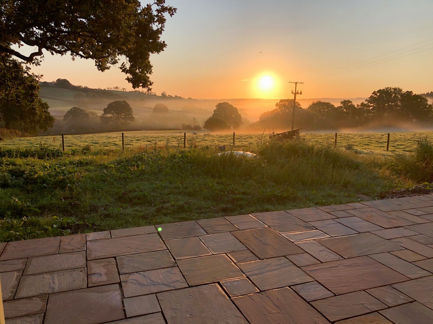 A patio with tiled flooring overlooking a foggy field with trees and a rising sun at The Cosy Cowshed in Dalwood