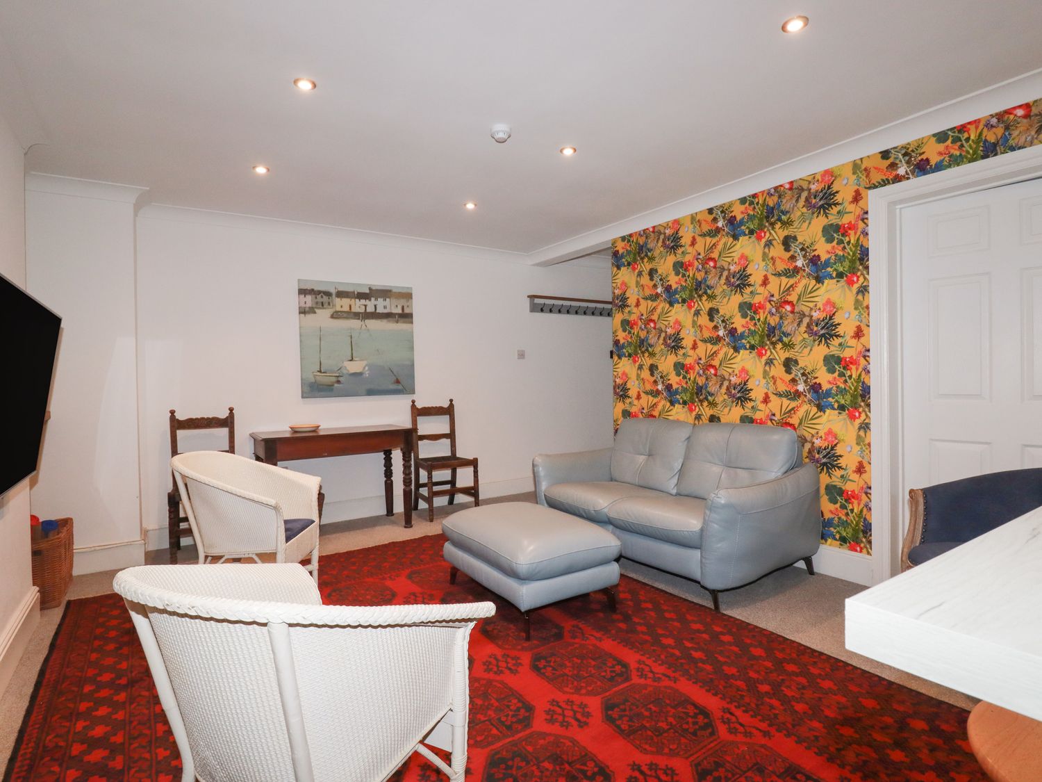 A living room with a gray sofa and ottoman two wicker chairs a red patterned rug a wooden table with two wooden chairs and a wall painting at Melvill Lower Deck in Falmouth