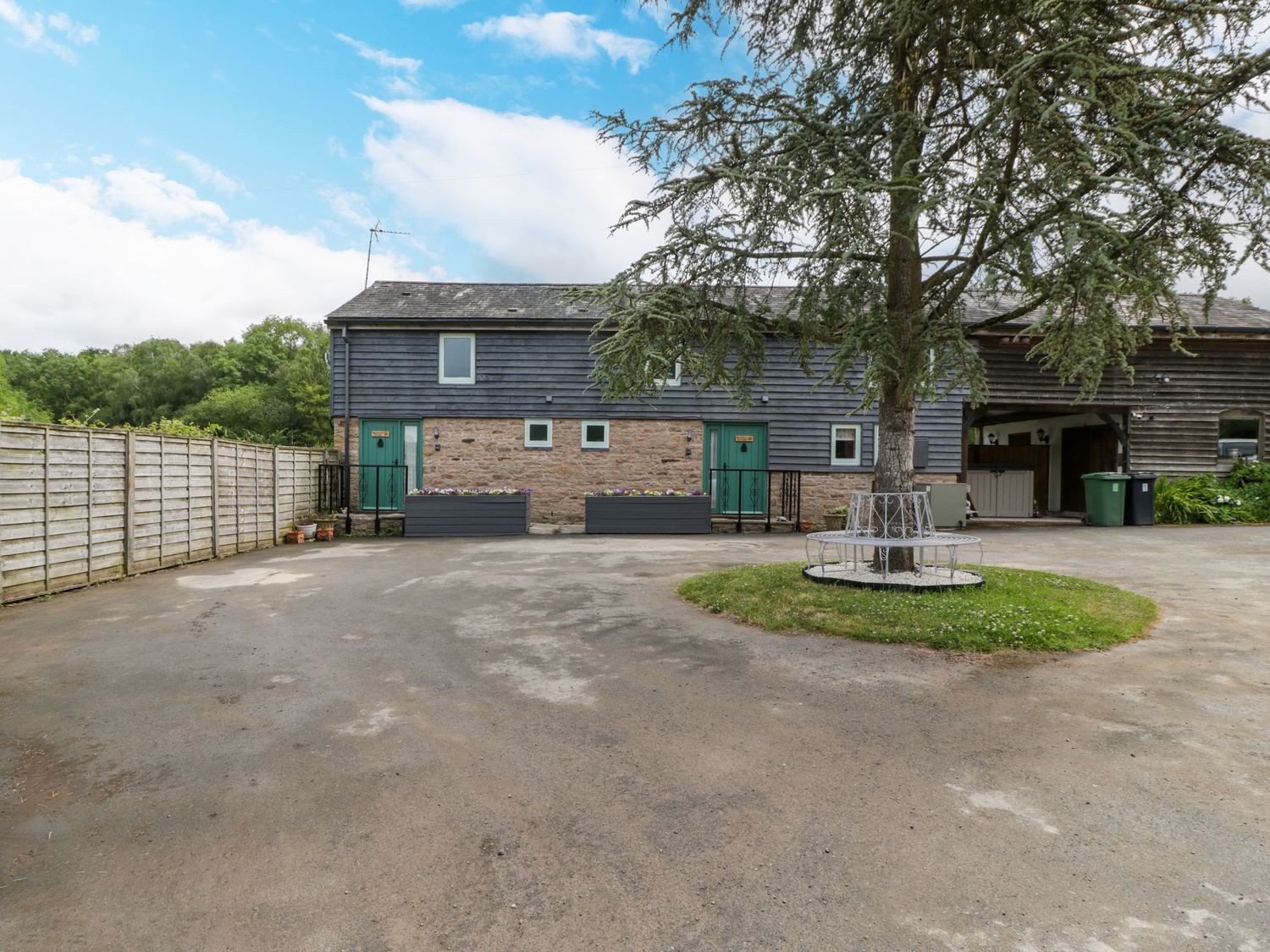 An outdoor view of a two-story building with green doors and stone and wood exterior with a tree in front surrounded by a circular bench at Beekeeper's Cottage in Pembridge