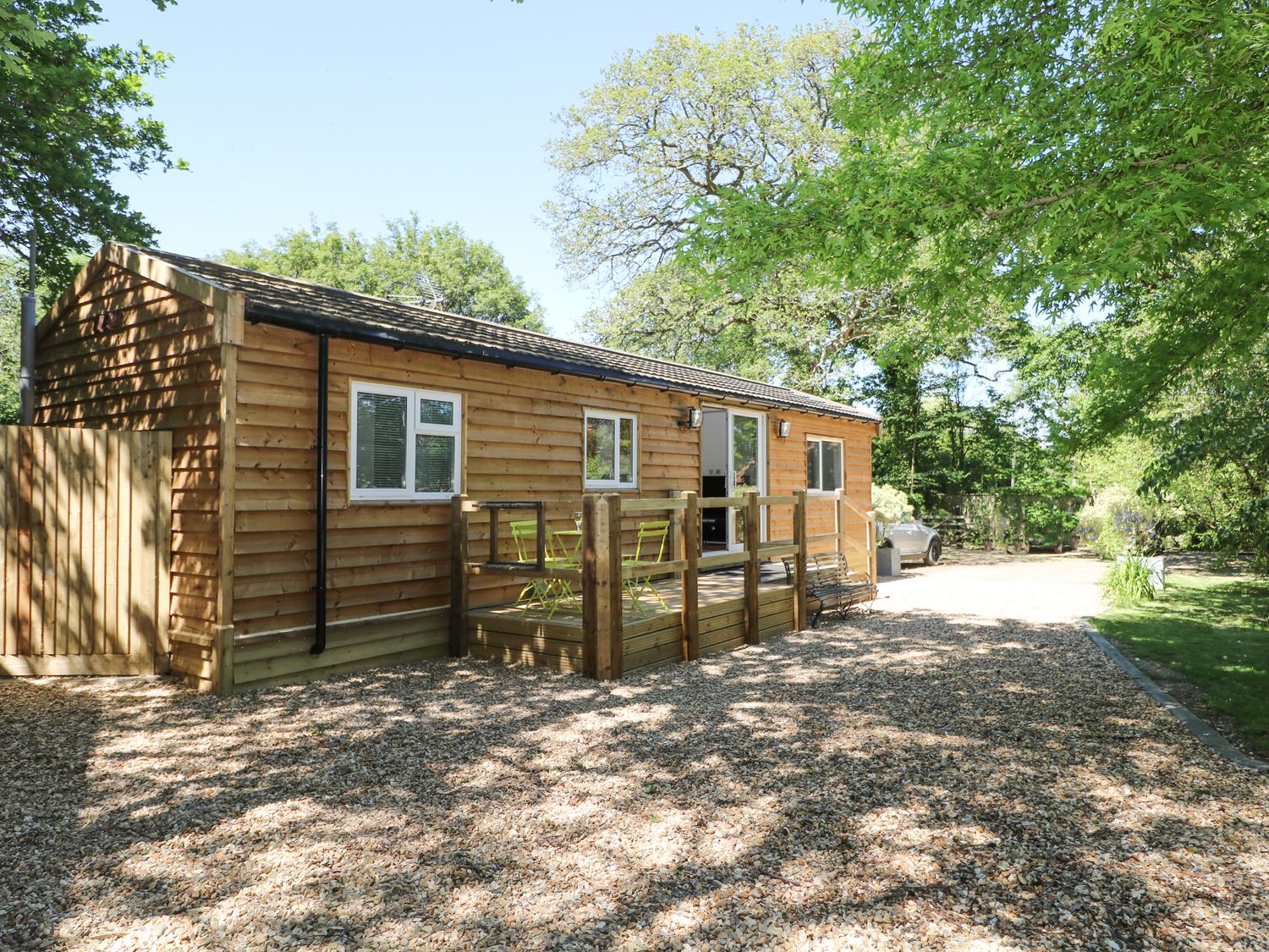 A wooden cabin with a small deck and green chairs surrounded by trees and a gravel driveway at Orchard Lodge in Bolney
