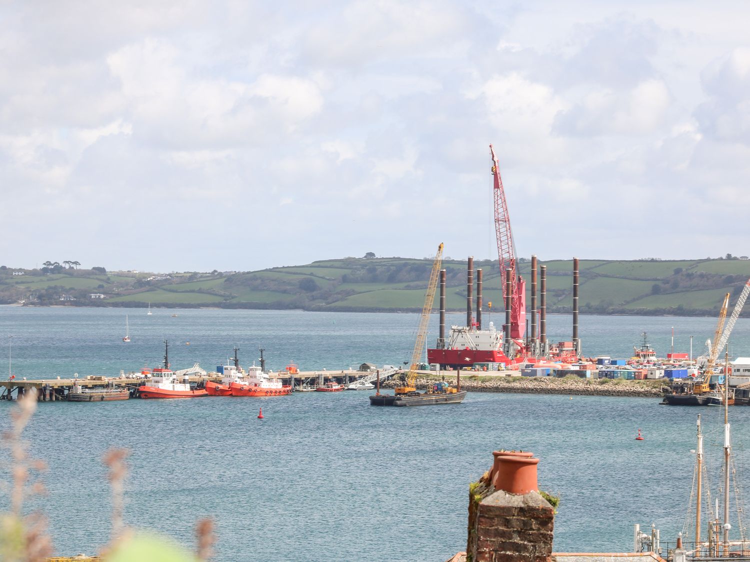 A harbor with boats cranes and a pier with green hills in the background at The Wesley Apartment in Falmouth