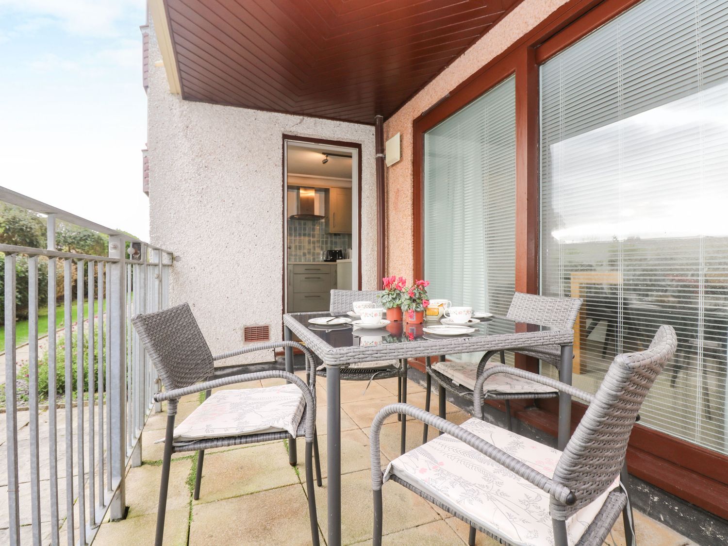 A balcony with a glass table and four wicker chairs with cushions and a kitchen visible through an open door at The Wesley Apartment in Falmouth