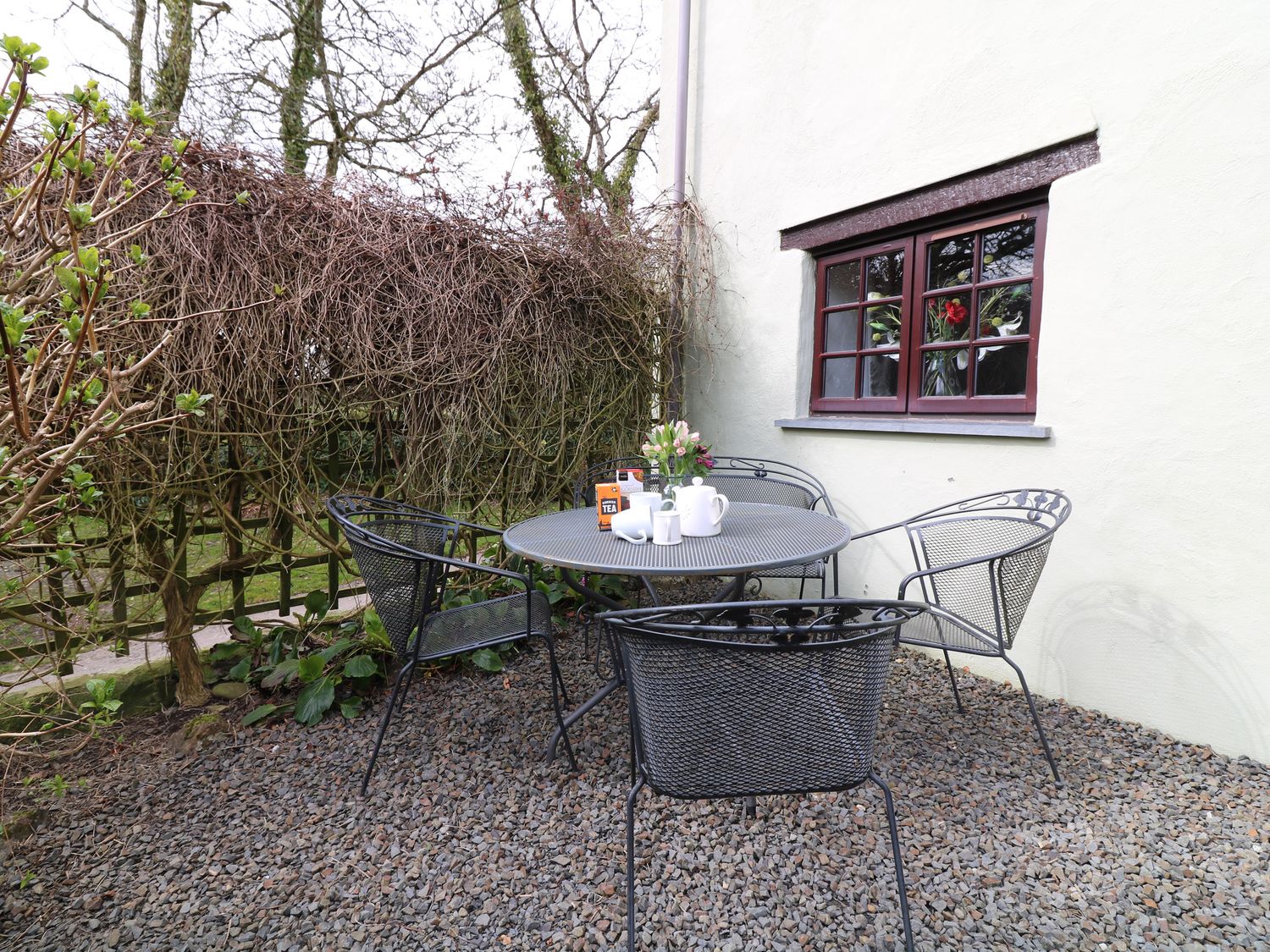 An outdoor seating area with a round metal table and four metal chairs on gravel near a house with a window and a trellis with bare vines at Beech Cottage Warbstow