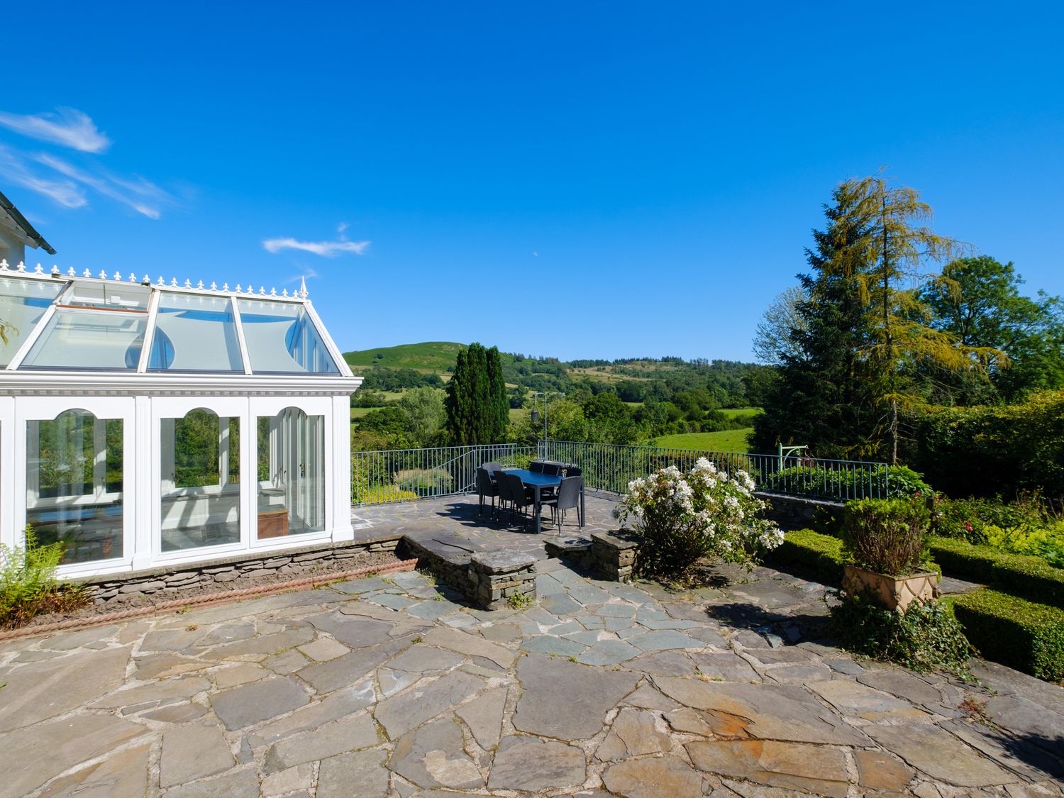 A stone patio with outdoor dining table and chairs beside a glass conservatory overlooking green hills and trees at Orchard Cottage in Outgate near Hawkshead