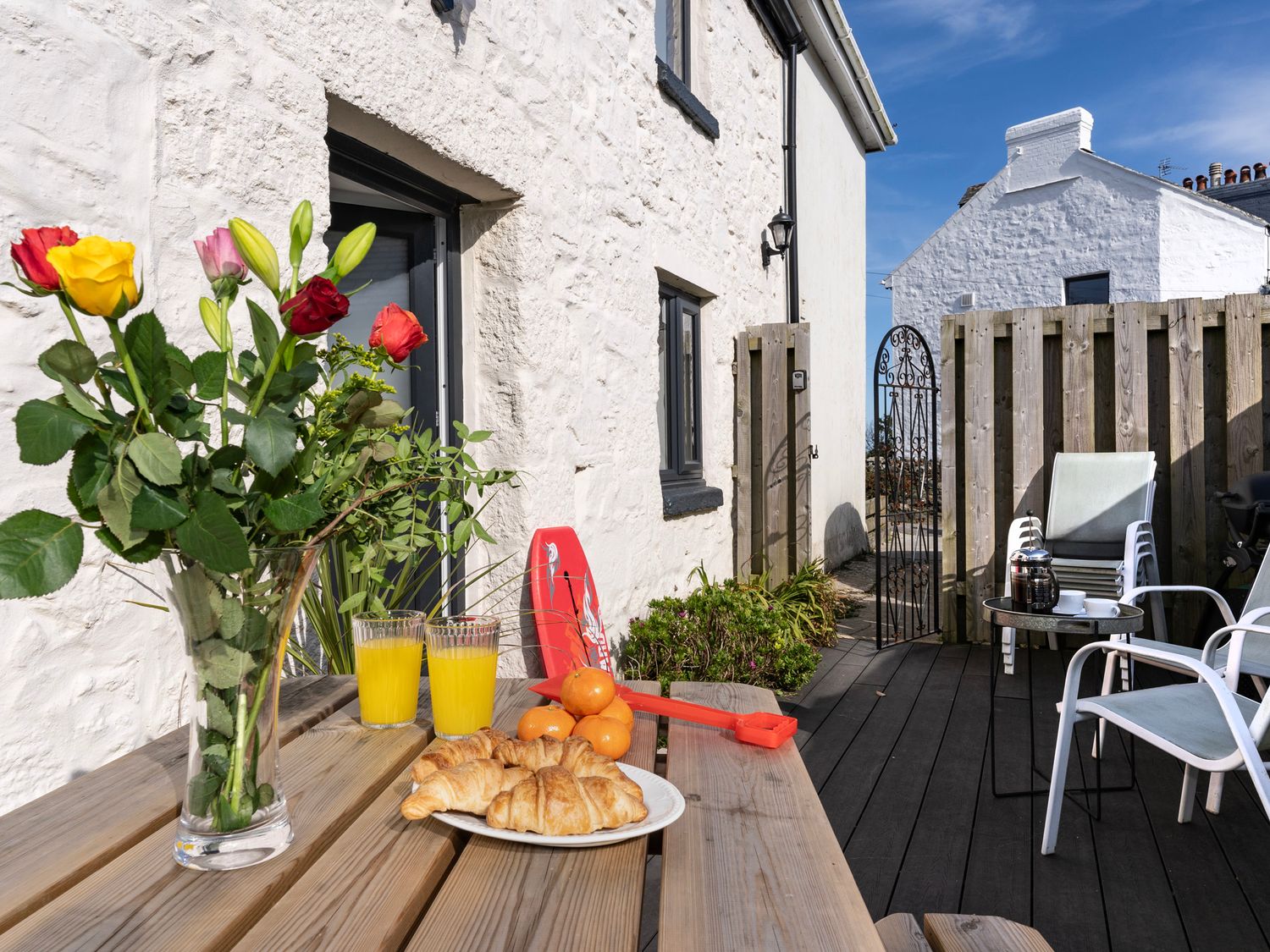 An outdoor patio with a wooden picnic table holding flowers croissants orange juice and mandarins at Rose Cottage in St Ives