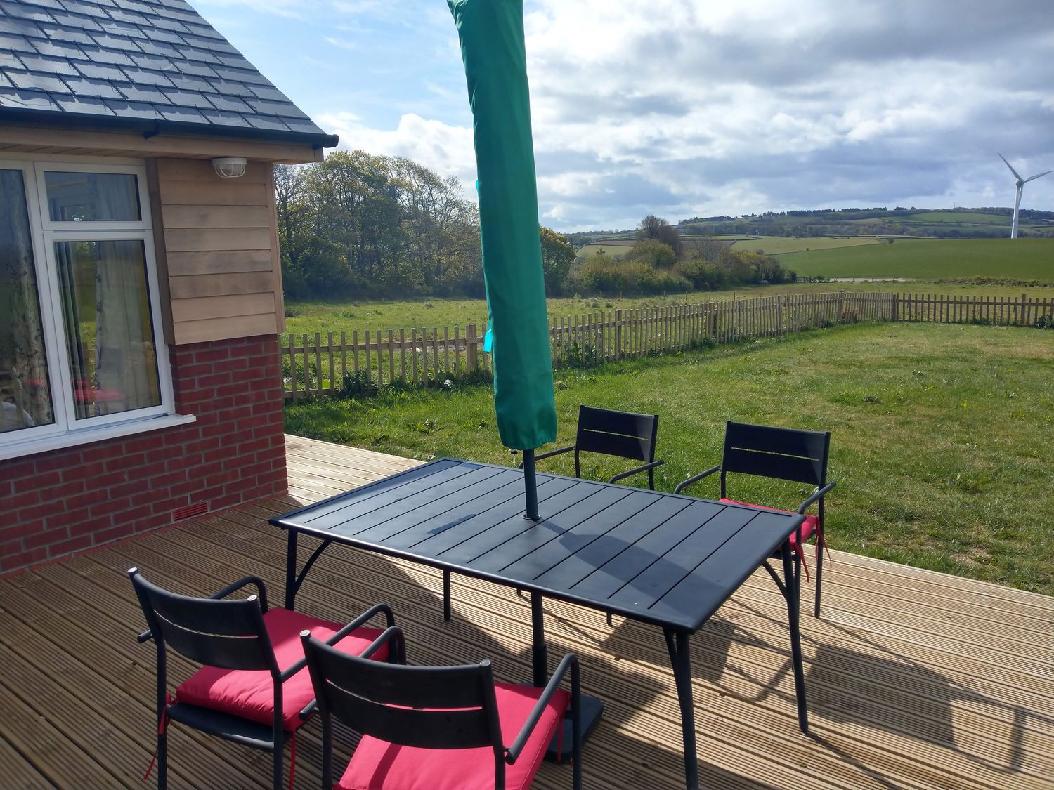A wooden deck with a black table and four black chairs with red cushions by a house with a fenced grassy yard and a wind turbine in the distance