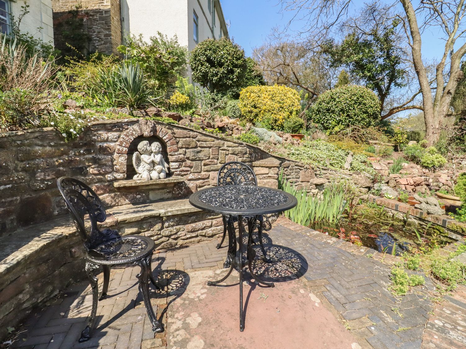A garden patio with black metal table and chairs and stone wall with cherub statue near a pond at The Carriage in Lydney