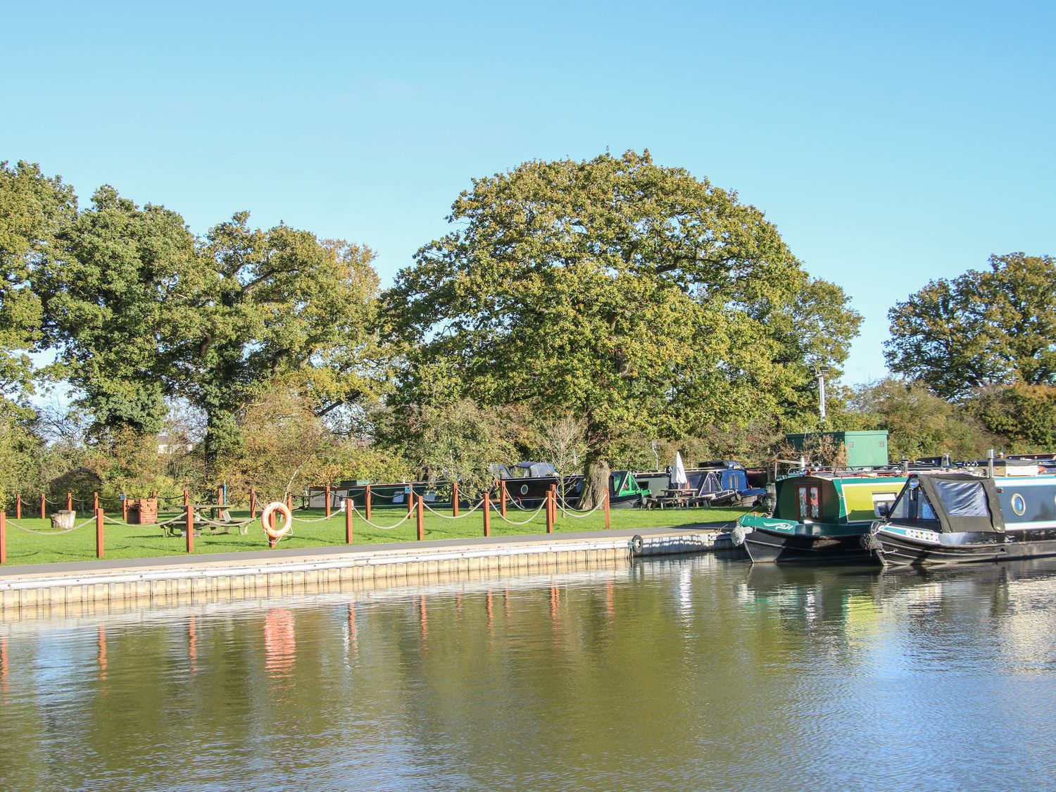 A canal with moored narrow boats and a grassy area with trees and picnic benches at The Tyre Barn in Whixall near Wem
