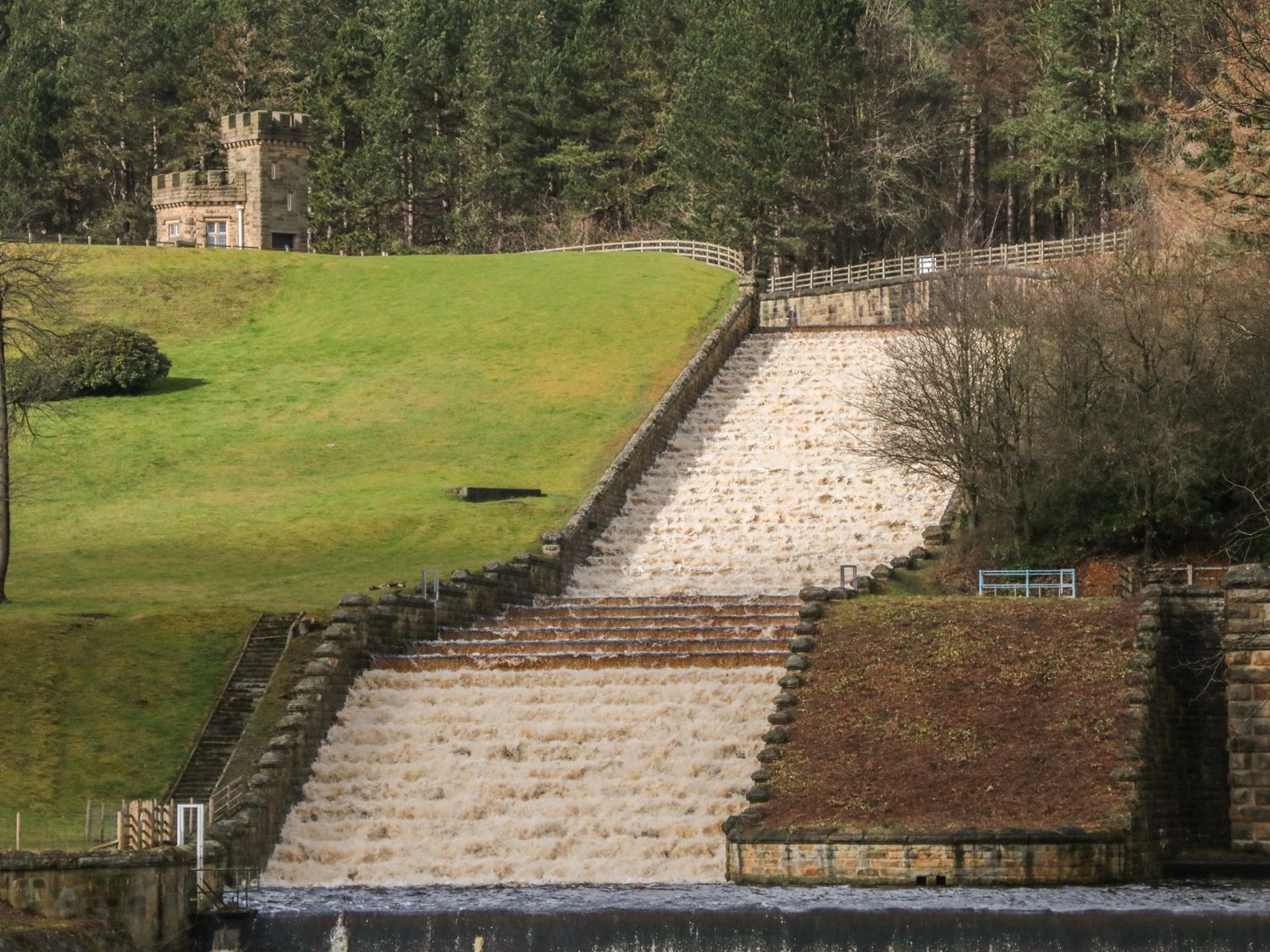 A cascading water spillway with grassy hills and trees surrounding a small stone tower at Chestnut Cottage in Stocksbridge