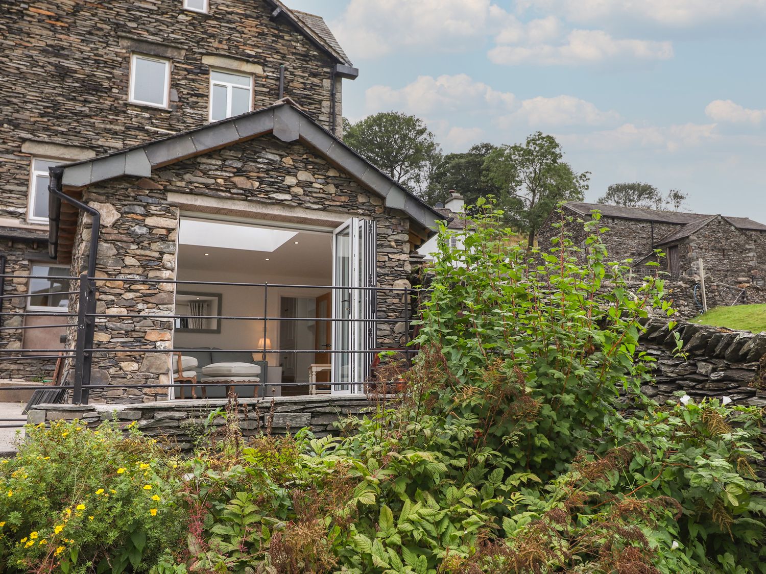 A stone house with large open doors leading to a room with chairs and a lamp surrounded by plants at Applethwaite View in Troutbeck near Troutbeck Bridge