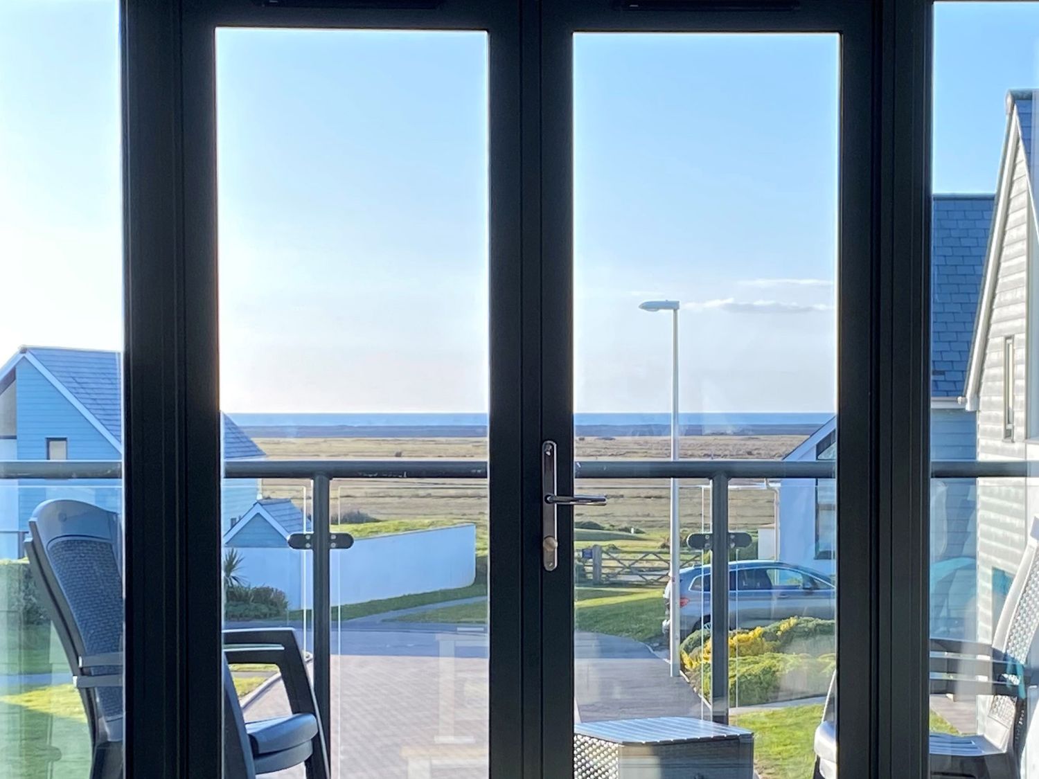 A view through glass doors of outdoor chairs a railing houses a street lamp and the sea beyond at Seaquel in Westward Ho