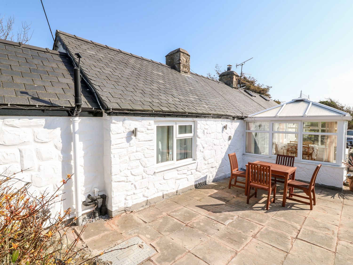 A white stone exterior cottage with a slate roof and a wooden outdoor table with chairs on a stone patio at Y Big in Morfa Nefyn