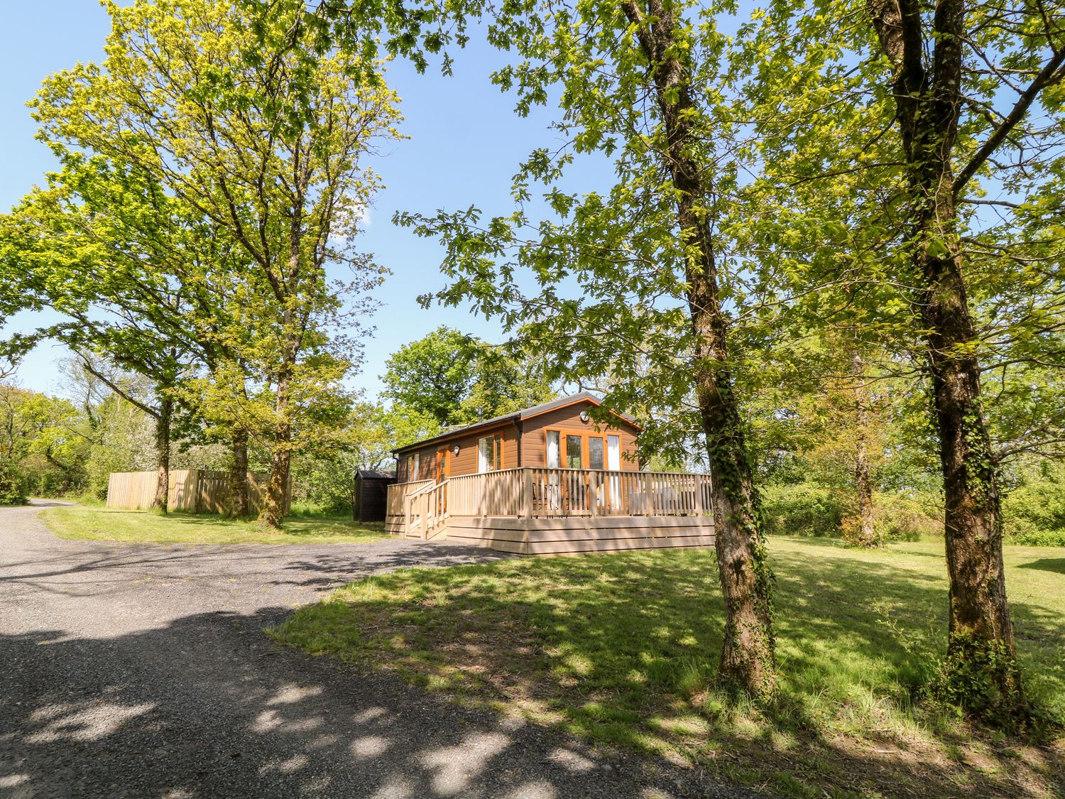 A wooden cabin with a porch surrounded by trees and grass at Dove Lodge in Highampton near Sheepwash