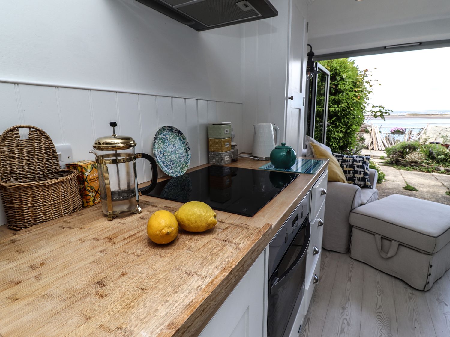 A kitchen counter with lemons a French press a wicker basket and a teapot next to a stove and oven with a sofa and an open door leading to a garden and water view at Mariners Cottage in Appledore