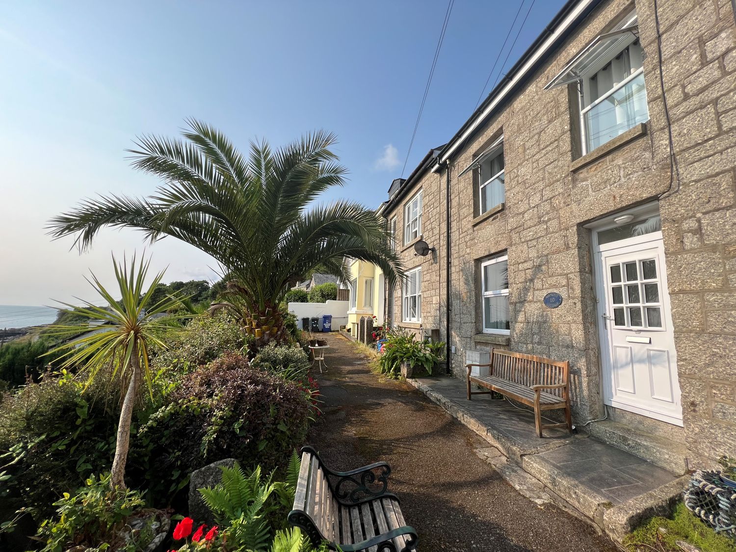 A stone house with white doors and windows beside a garden with palm trees and benches at Lookout Cottage in Newlyn