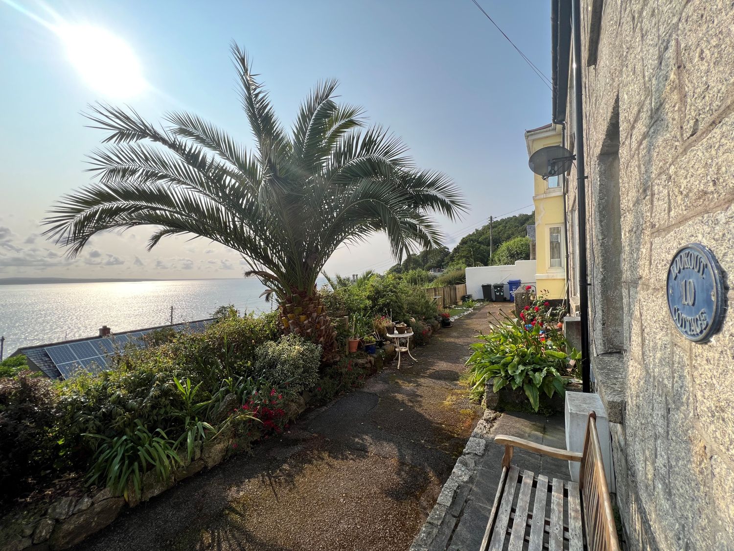A pathway with plants and a bench next to a stone building with a palm tree by the sea at Lookout Cottage in Newlyn