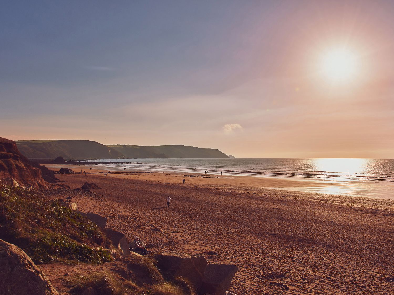 A beach with people walking near the water and rocky cliffs in the background at The Linhay in Holsworthy