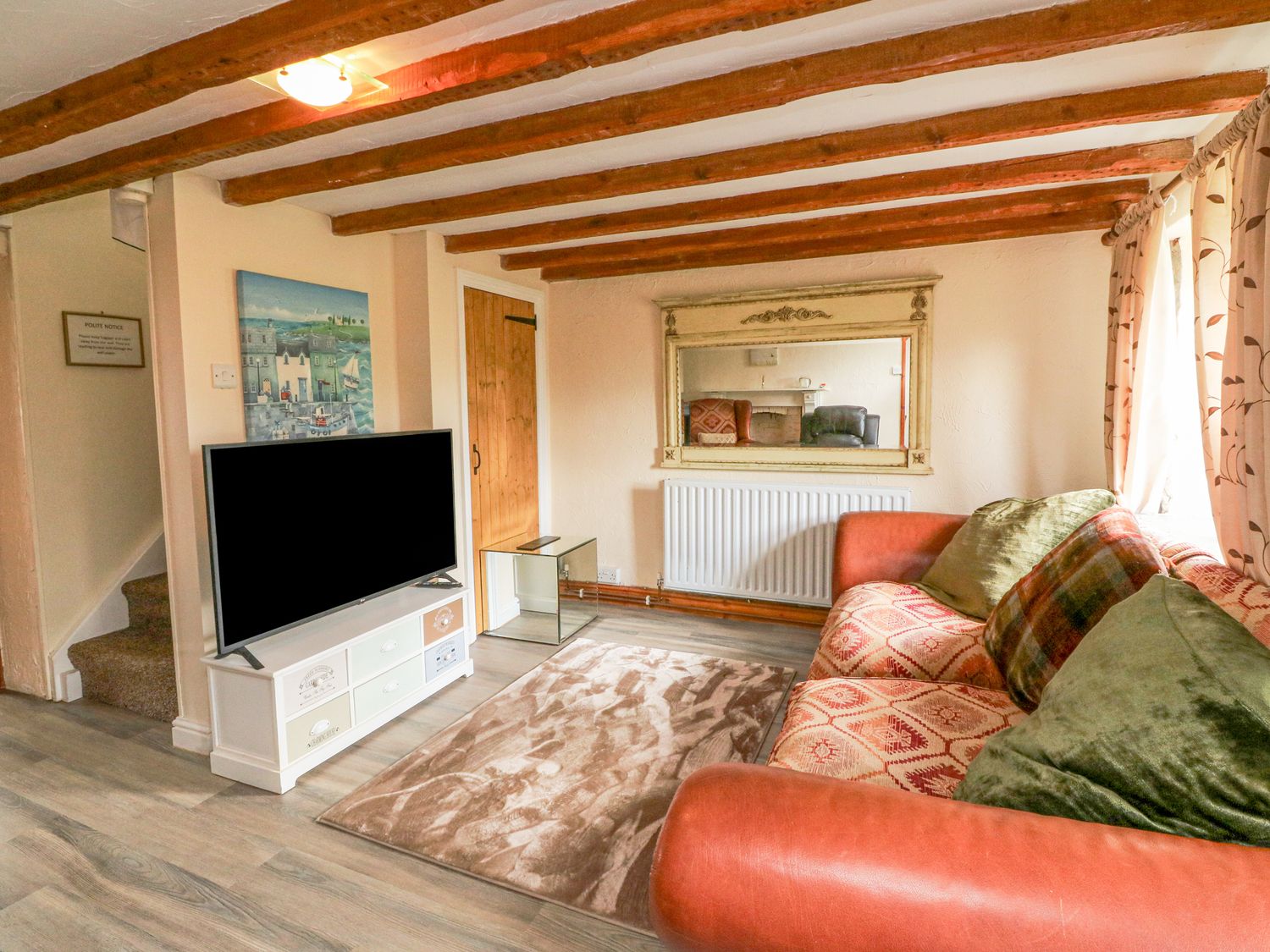 A living room with a patterned sofa and cushions a wall mirror a television on a stand and wooden ceiling beams at The Linhay in Holsworthy