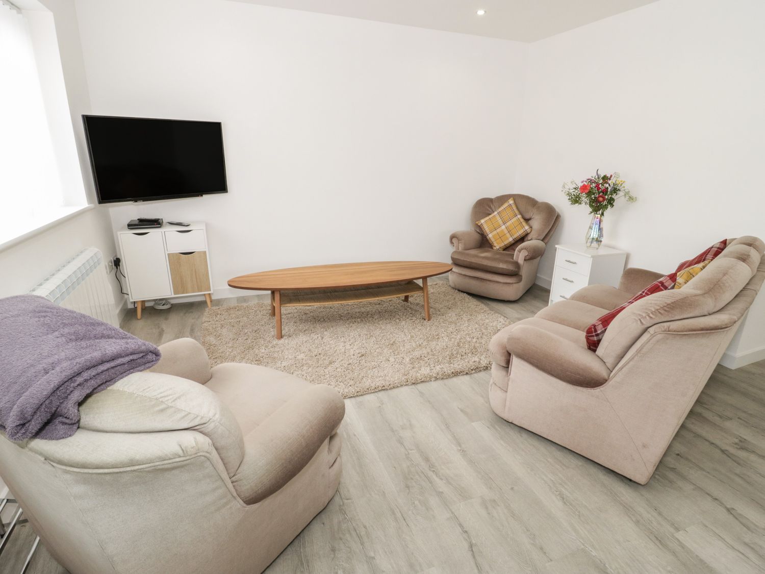 A living room with beige armchairs a wooden coffee table a wall mounted television and a small white cabinet with flowers on a side table at Trigfa in Llanfechell
