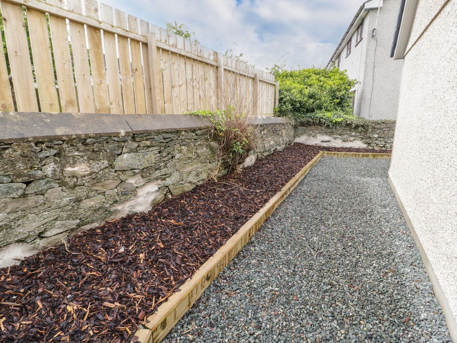 An outdoor area with a gravel path beside a white textured building and a wooden raised bed with mulch along a stone wall and wooden fence at Trigfa in Llanfechell