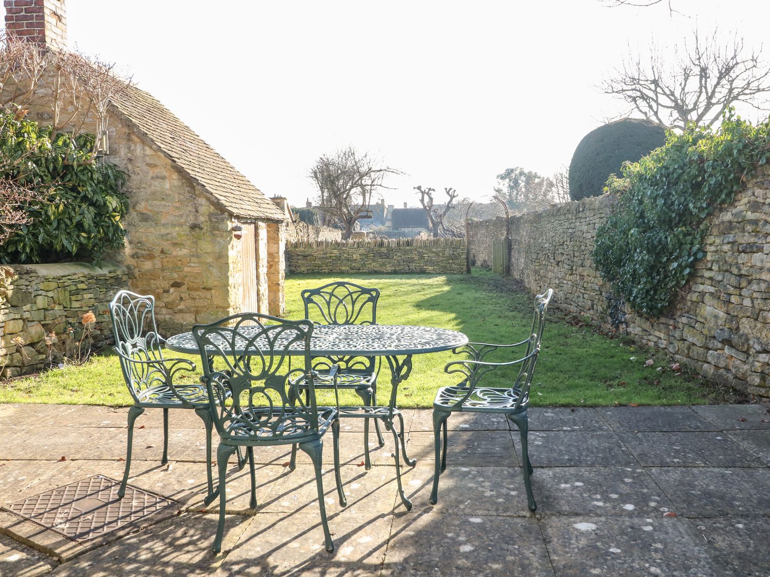 A stone patio with a metal table and four chairs next to a stone building and garden at Cowfair Cottage in Chipping Campden