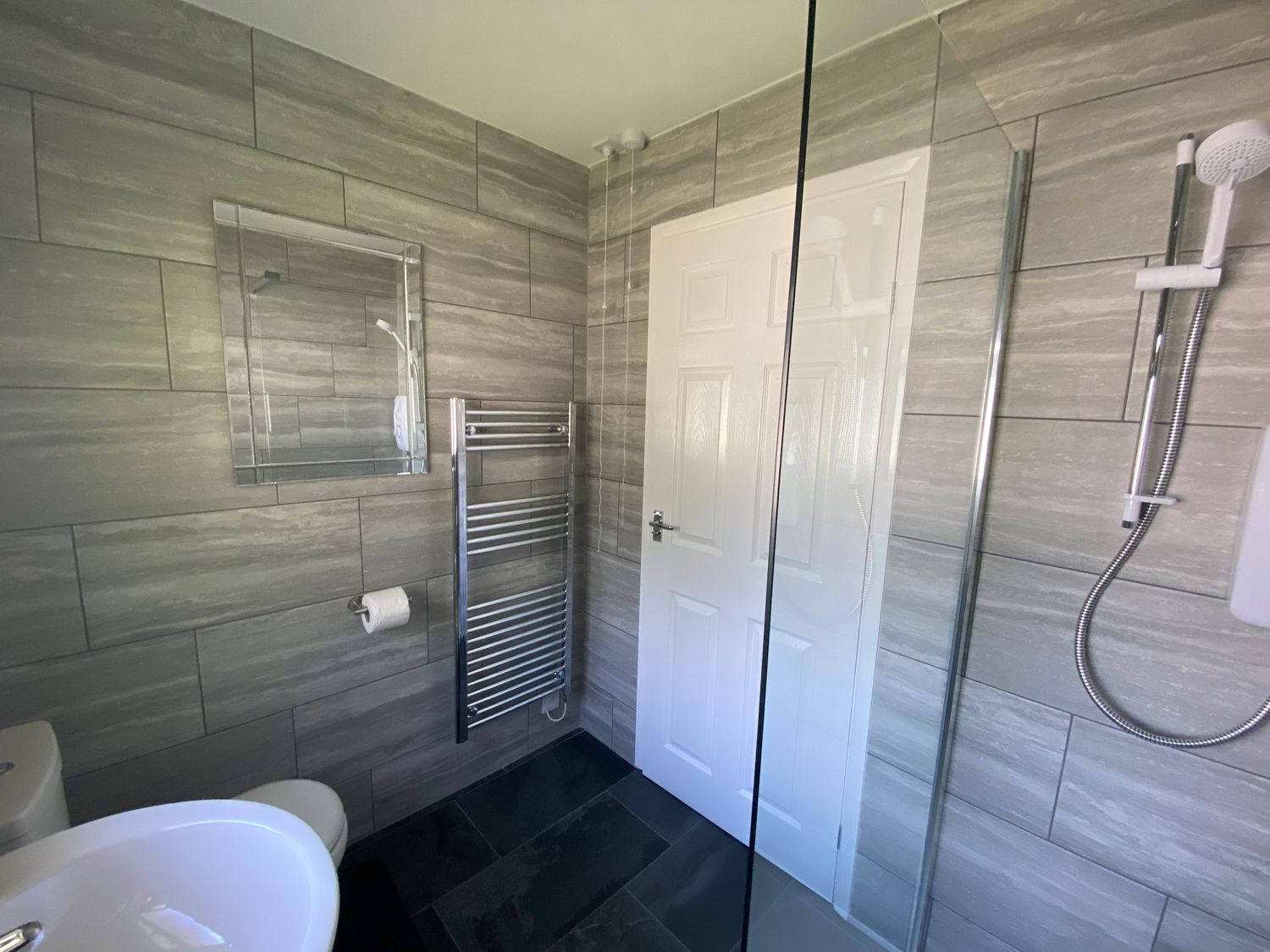 A bathroom with a sink, toilet, wall mirror, towel radiator, showerhead, and a white door at Lands End in Liskeard near St Cleer