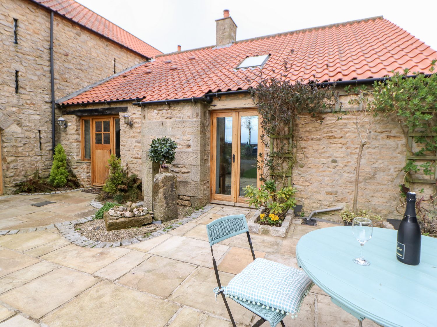 A stone house with a red tiled roof and wooden doors a small patio with a blue table and chair with a cushion a champagne bottle and glass on the table at Crakesmire House in Headlam near Gainford