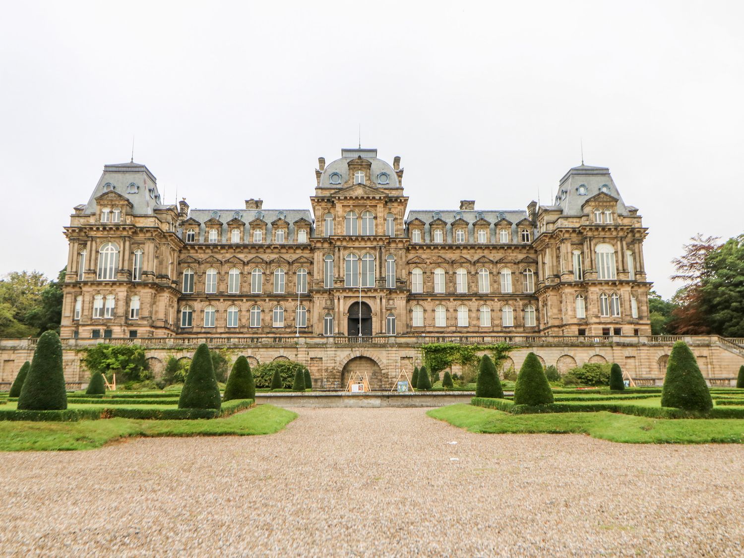 A large historic building with many windows and mansard roofs in front of a formal garden with cone-shaped shrubs and gravel paths at Crakesmire House Headlam near Gainford